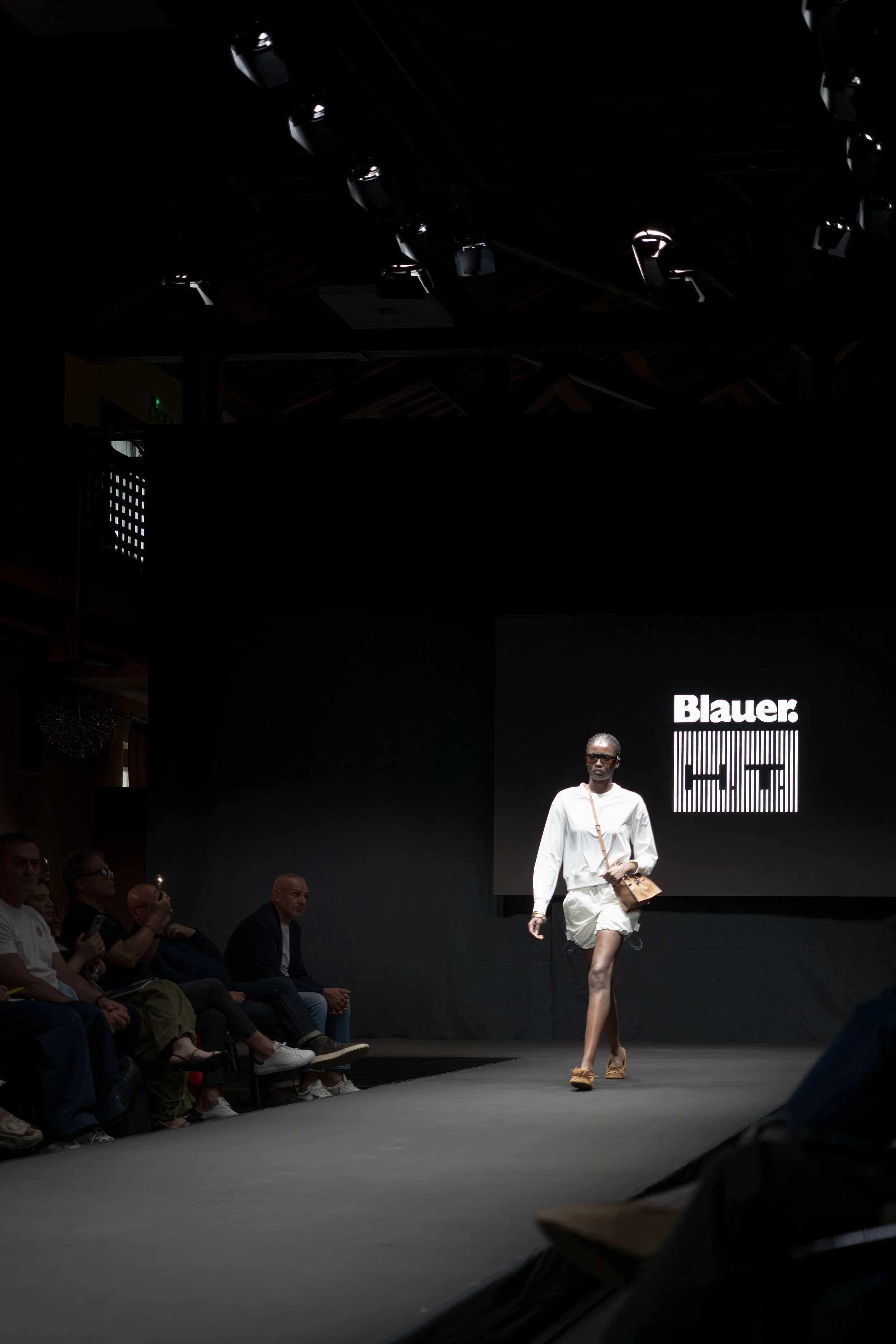 Una modella cammina sulla passerella durante una sfilata di moda, con un pubblico seduto ai lati. Sullo sfondo si vede un grande schermo con il logo 'Blauer'.