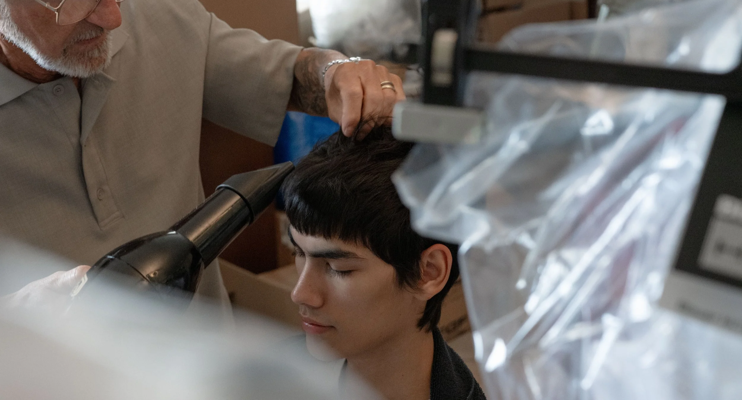 Un uomo anziano con barba che fa un trattamento di tricotomia a un ragazzo con capelli corti e scuri. Sono in un ambiente clinico o medico, con attrezzature di sterilizzazione e materiali chirurgici visibili.