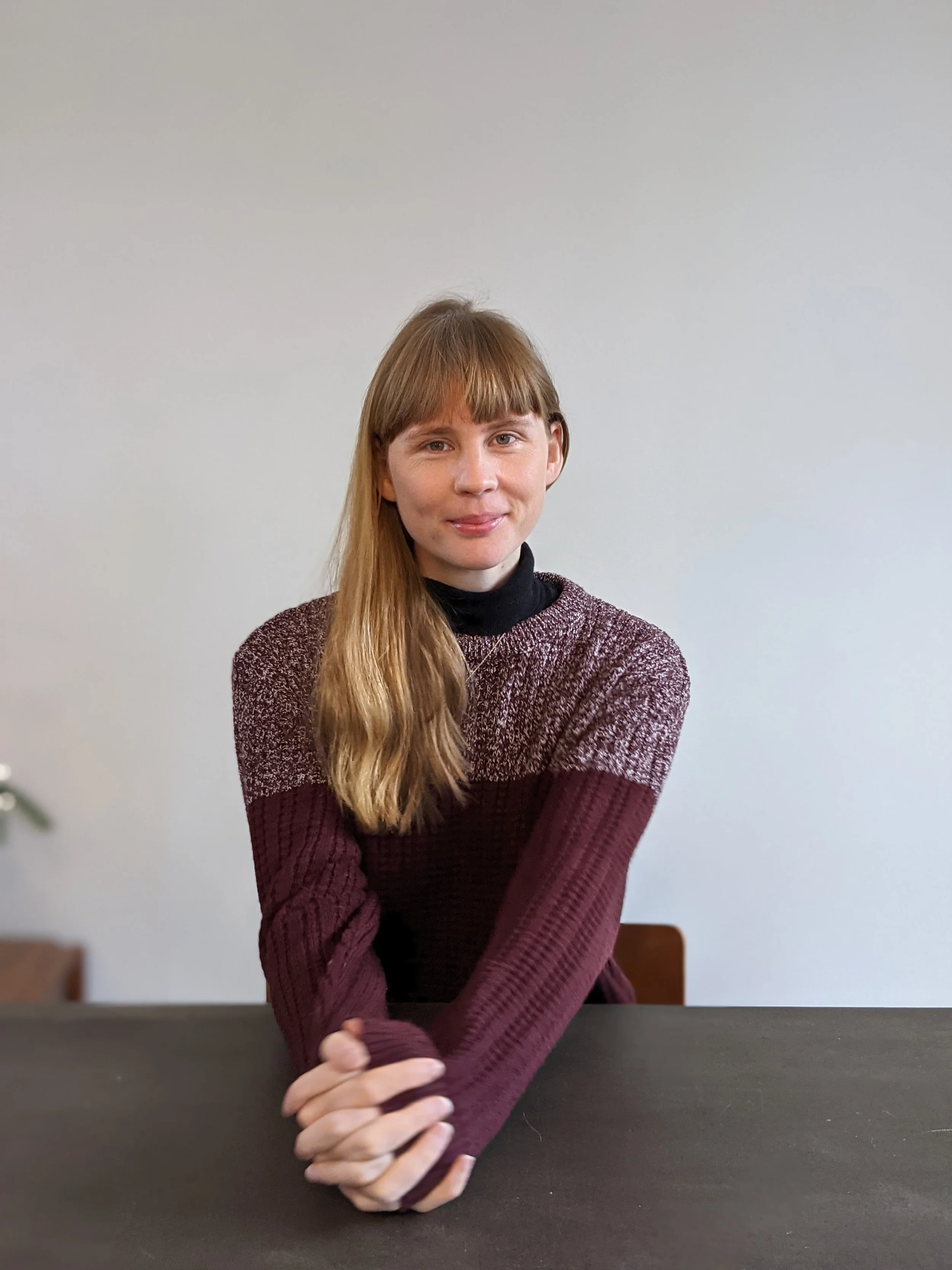 A young woman with long blonde hair and bangs wearing a maroon and black sweater, sitting at a table against a plain, light-colored wall, with her hands clasped on the table.