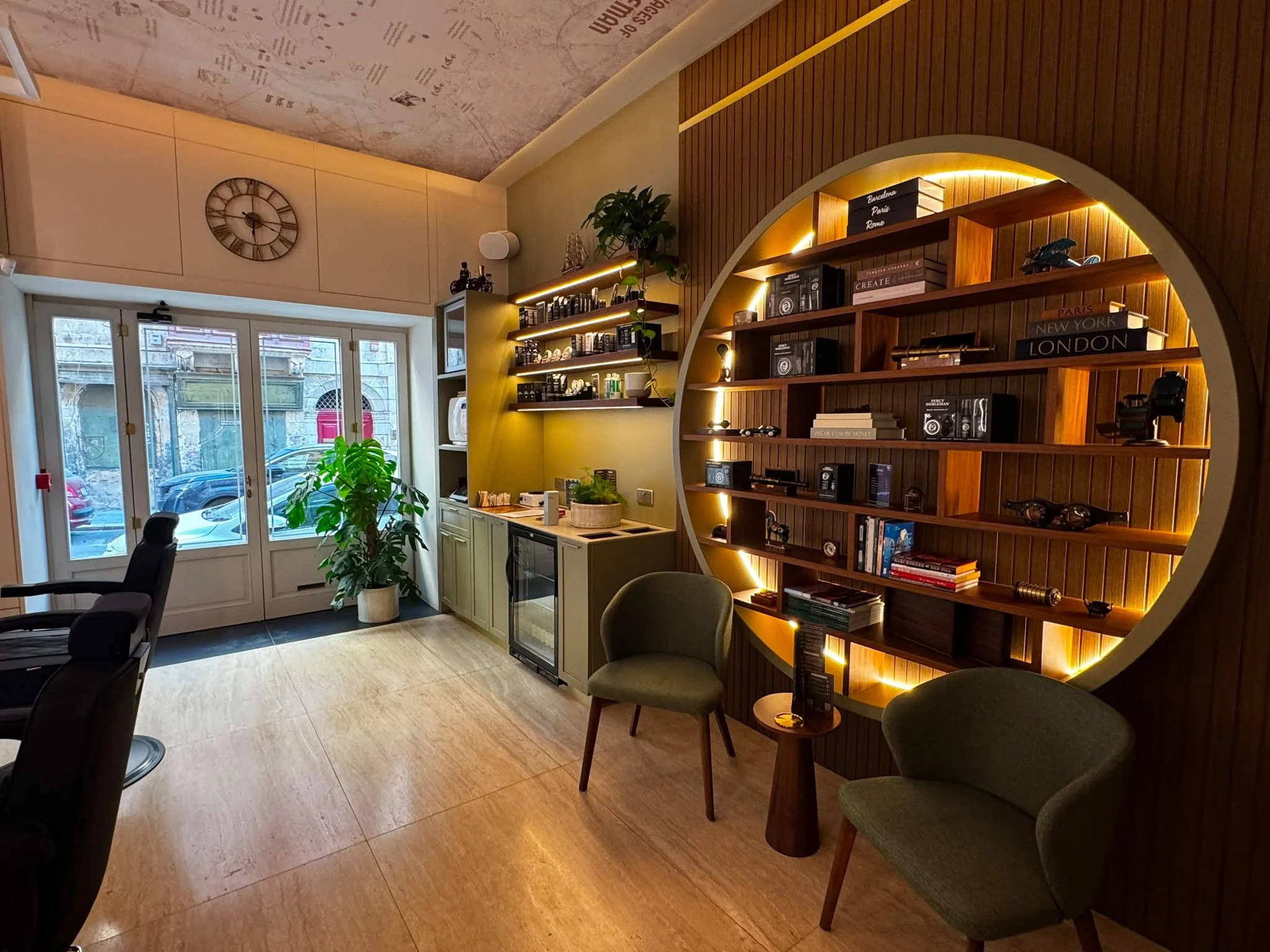 Cozy interior of a Percy Nobleman barbershop with large window, wooden bookshelves, green chairs, and decorative lighting.