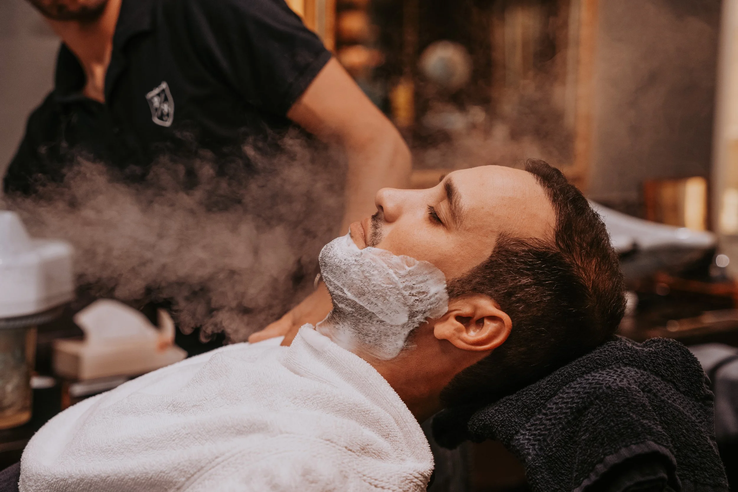 A man is receiving a shave at Percy Nobleman Barbershop as white shaving foam is applied to his face.