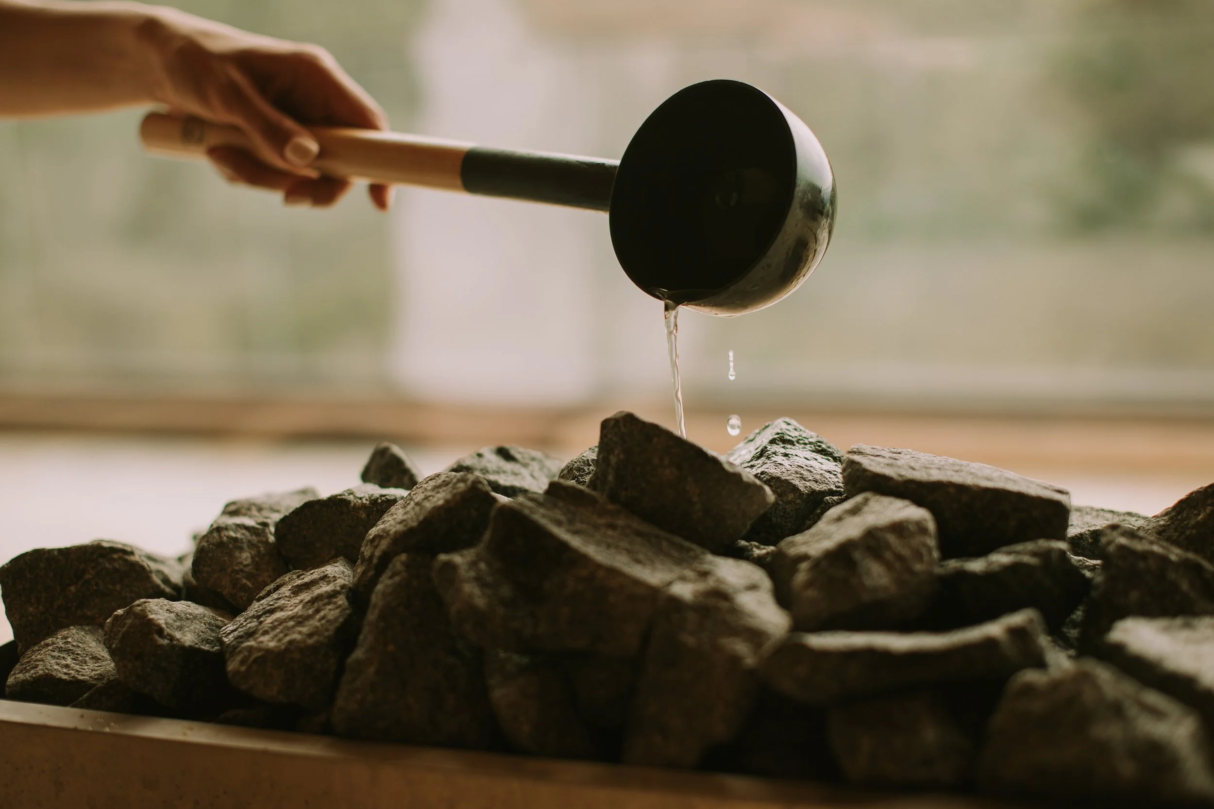 A hand holding a black ladle pouring liquid over rocks or stones.