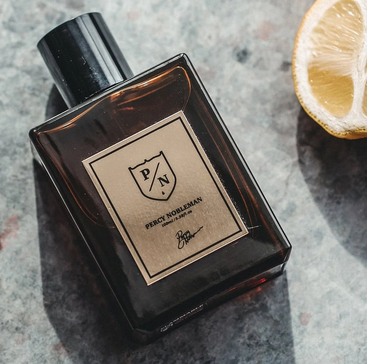 A bottle of Percy Nobleman fragrance product with a black cap, positioned on a gray surface beside a lemon wedge.
