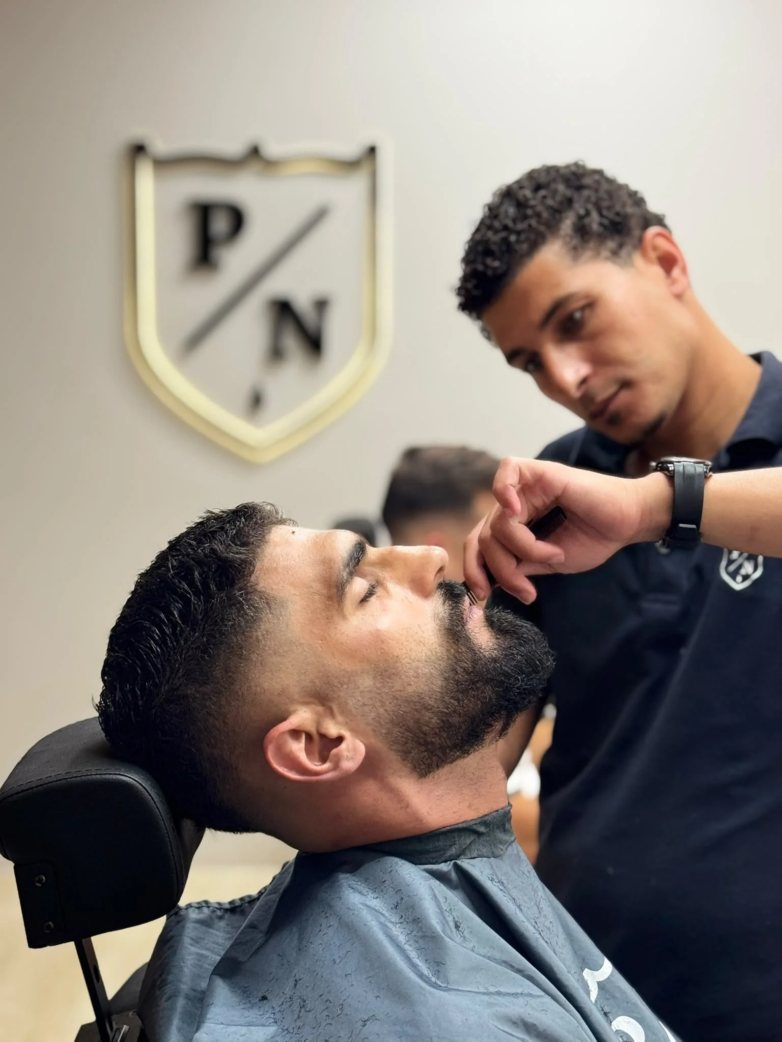 A man with dark hair and beard sitting in a barber's chair, getting a shave from a barber