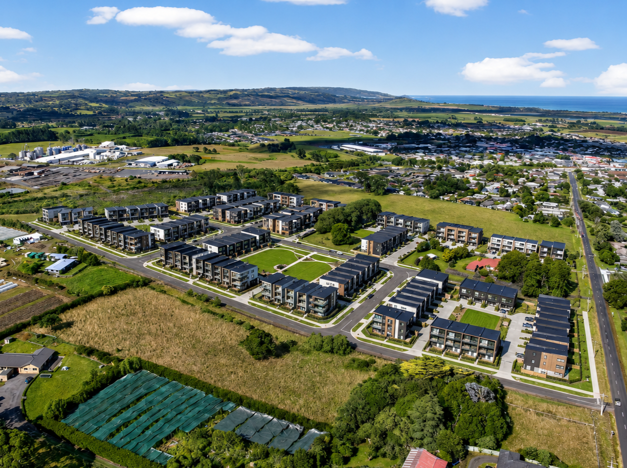 Aerial view of a large green field with a red boundary outline, including a small house with a red roof, surrounded by trees and neighboring houses, with a town and hills in the background under a partly cloudy sky.