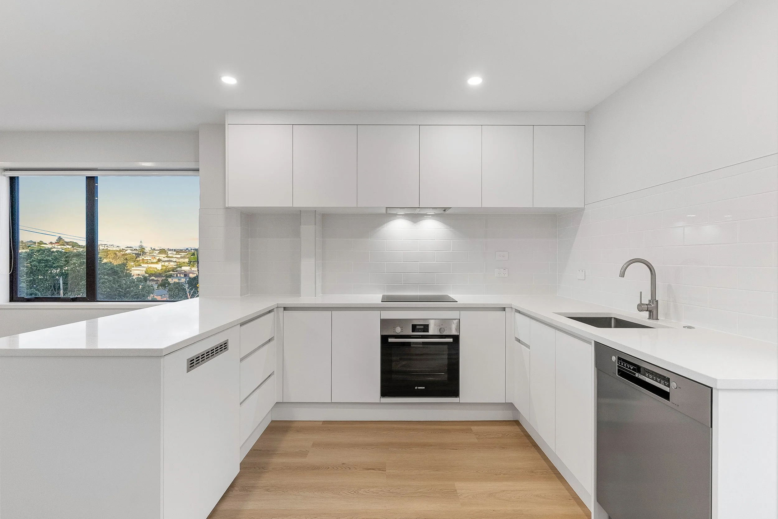 Modern white kitchen with built-in appliances, countertops, and a large window showing a cityscape view.