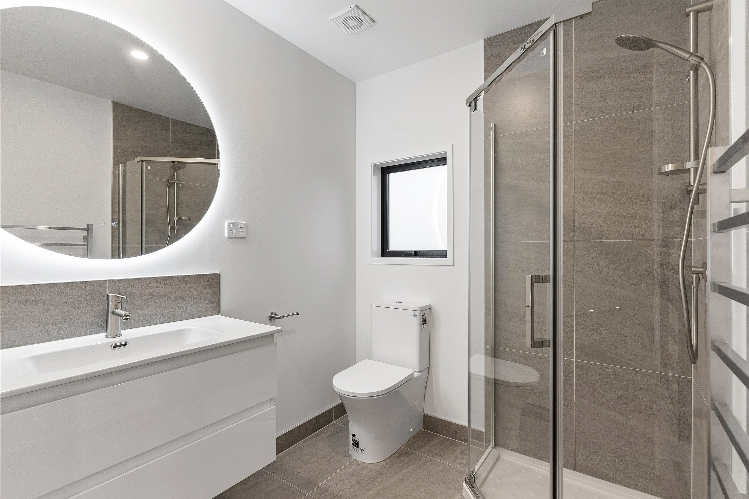 Modern bathroom with a white vanity, a round mirror with backlighting, a toilet, a small window, and a glass-enclosed shower with gray tiles.