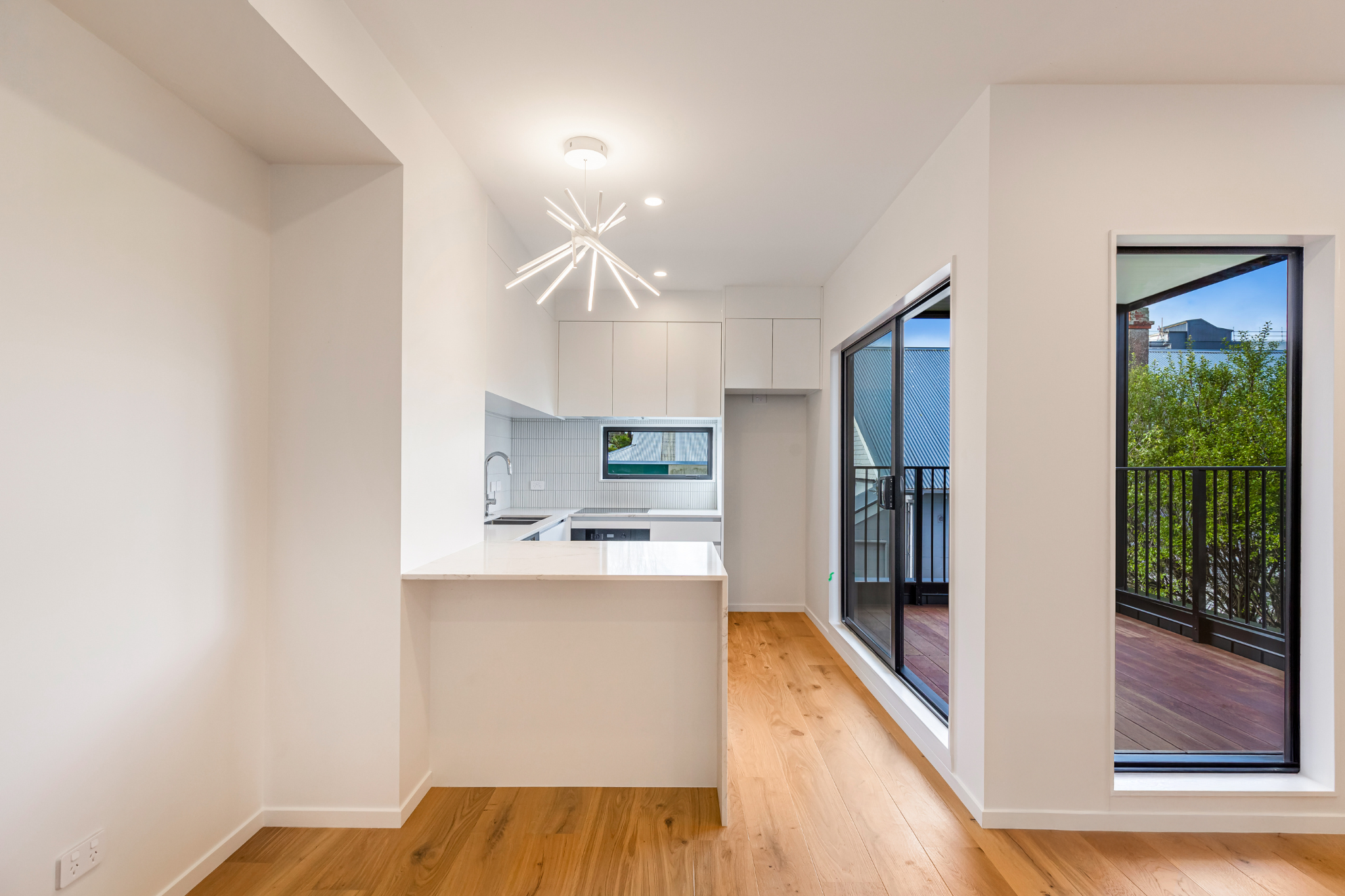 Bright modern kitchen with white cabinets, a small window, and a sliding glass door leading to an outdoor balcony with black railing, hardwood floors, and a minimalist white ceiling light fixture.