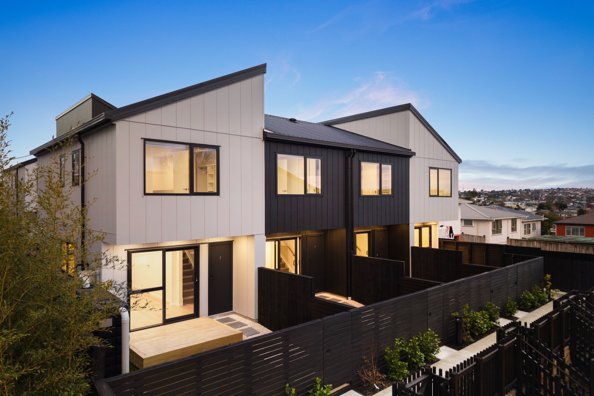 Modern two-story residential building with white and black exterior panels, large windows, a small outdoor deck, and a black fence, during evening with lights on inside.