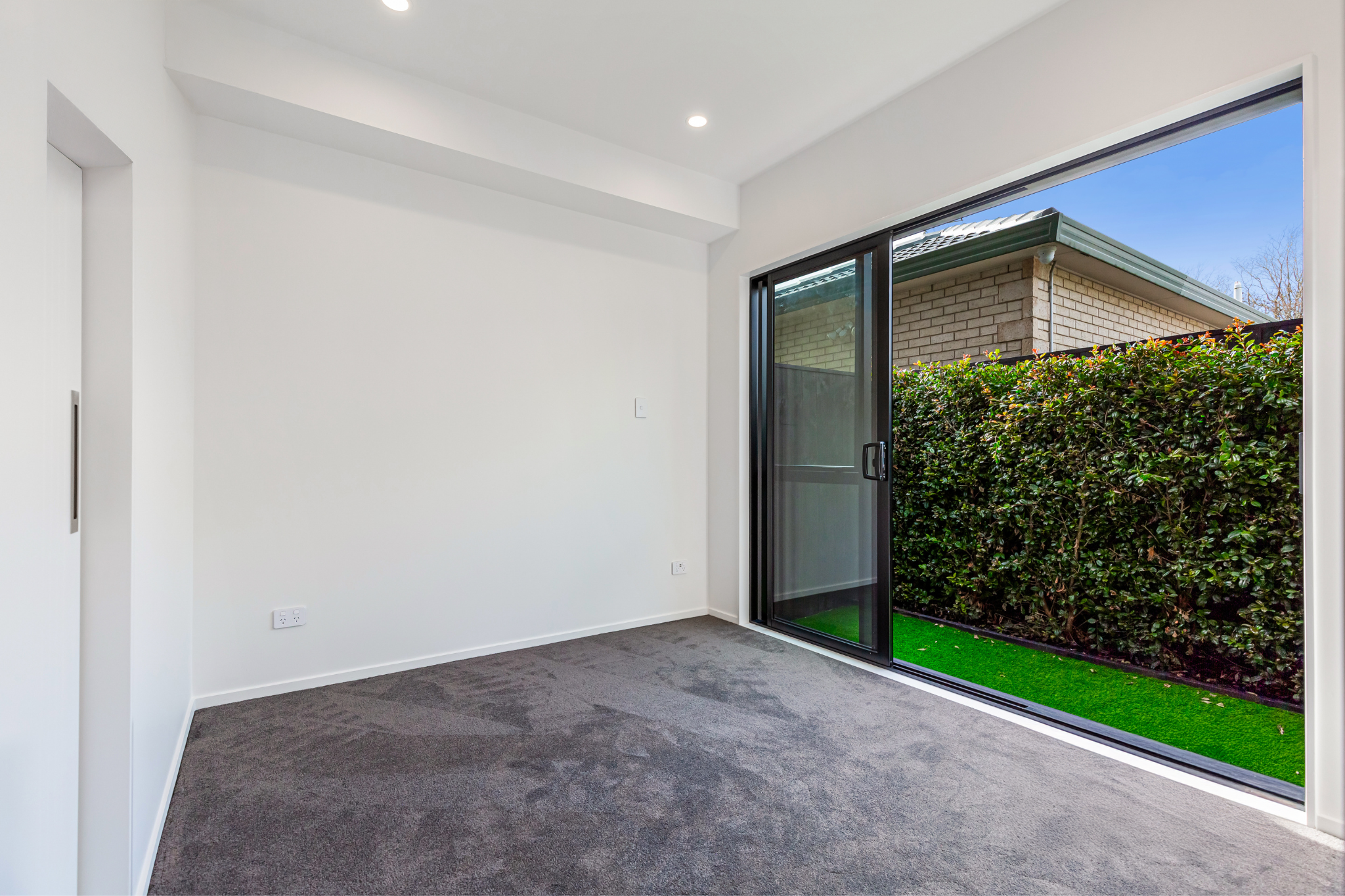 Empty room with gray carpet, white walls, and a large glass sliding door opening to a small backyard with green grass and bushes.
