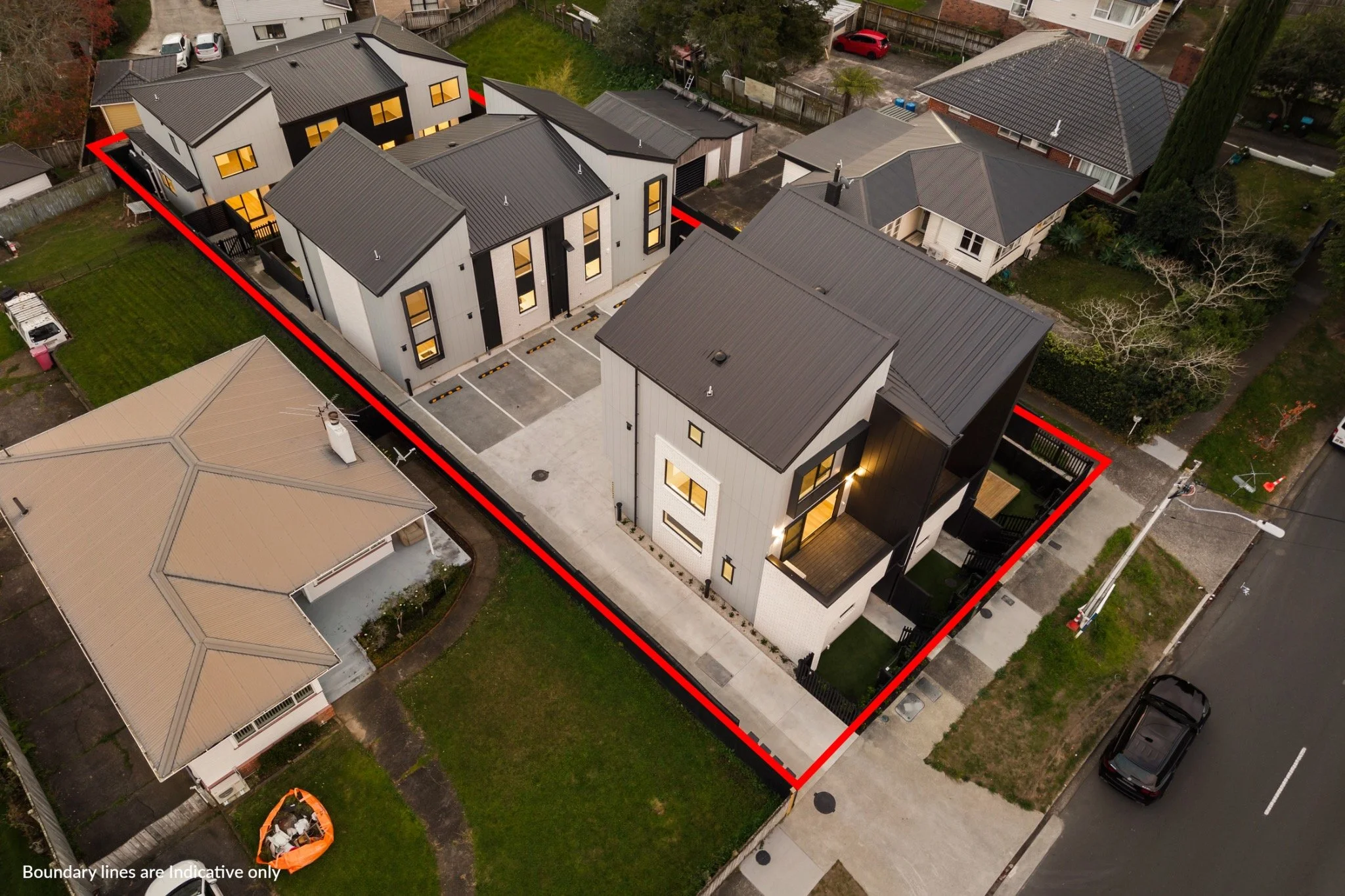 An aerial view of a modern residential property with multiple white and black buildings, a small backyard, and parking spaces. Red boundary lines outline the property borders.