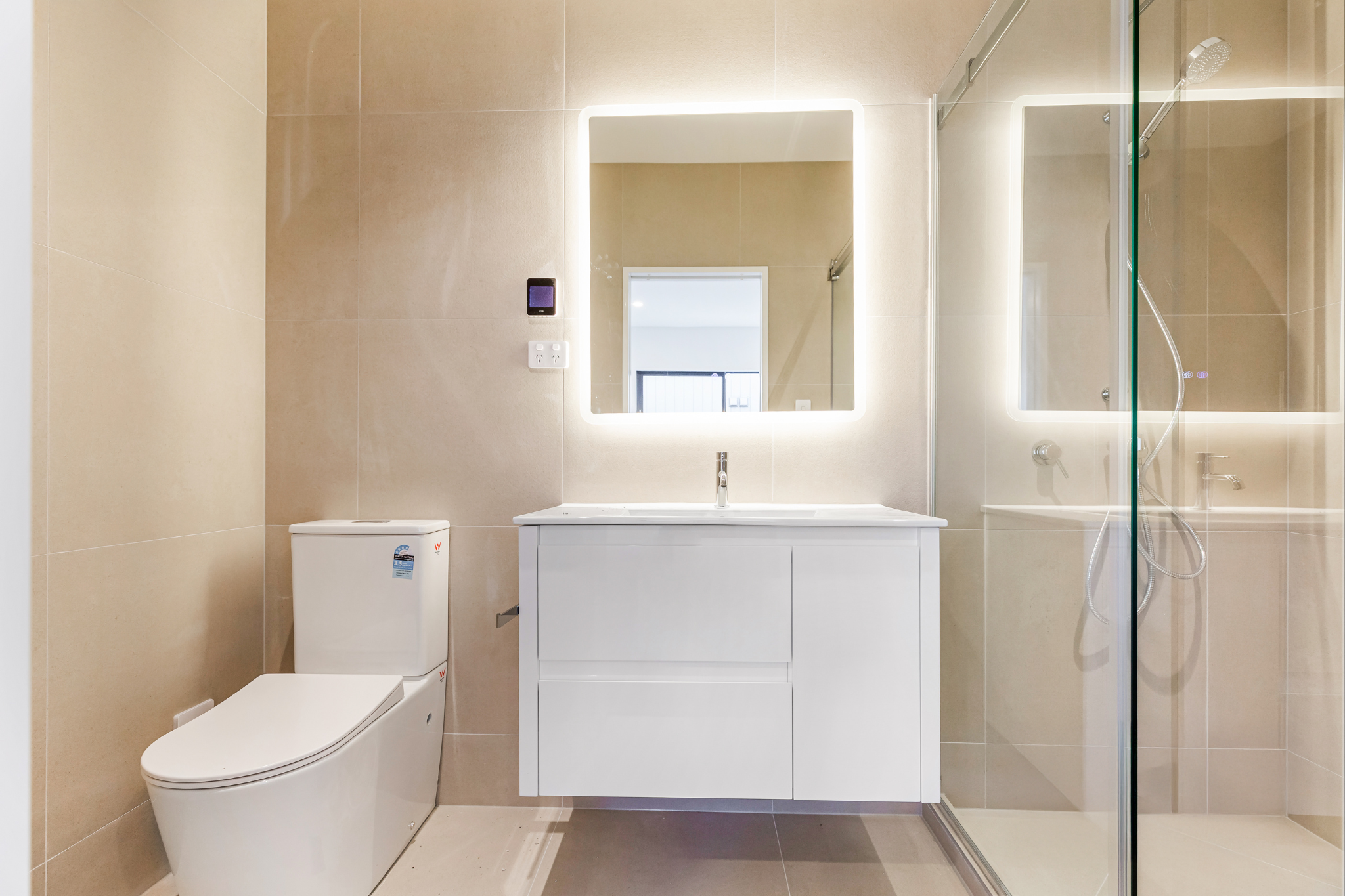 Modern bathroom with beige tiled walls and floor, a white toilet, a white vanity with a sink and mirror with built-in LED lights, and a glass-enclosed shower with a handheld showerhead.