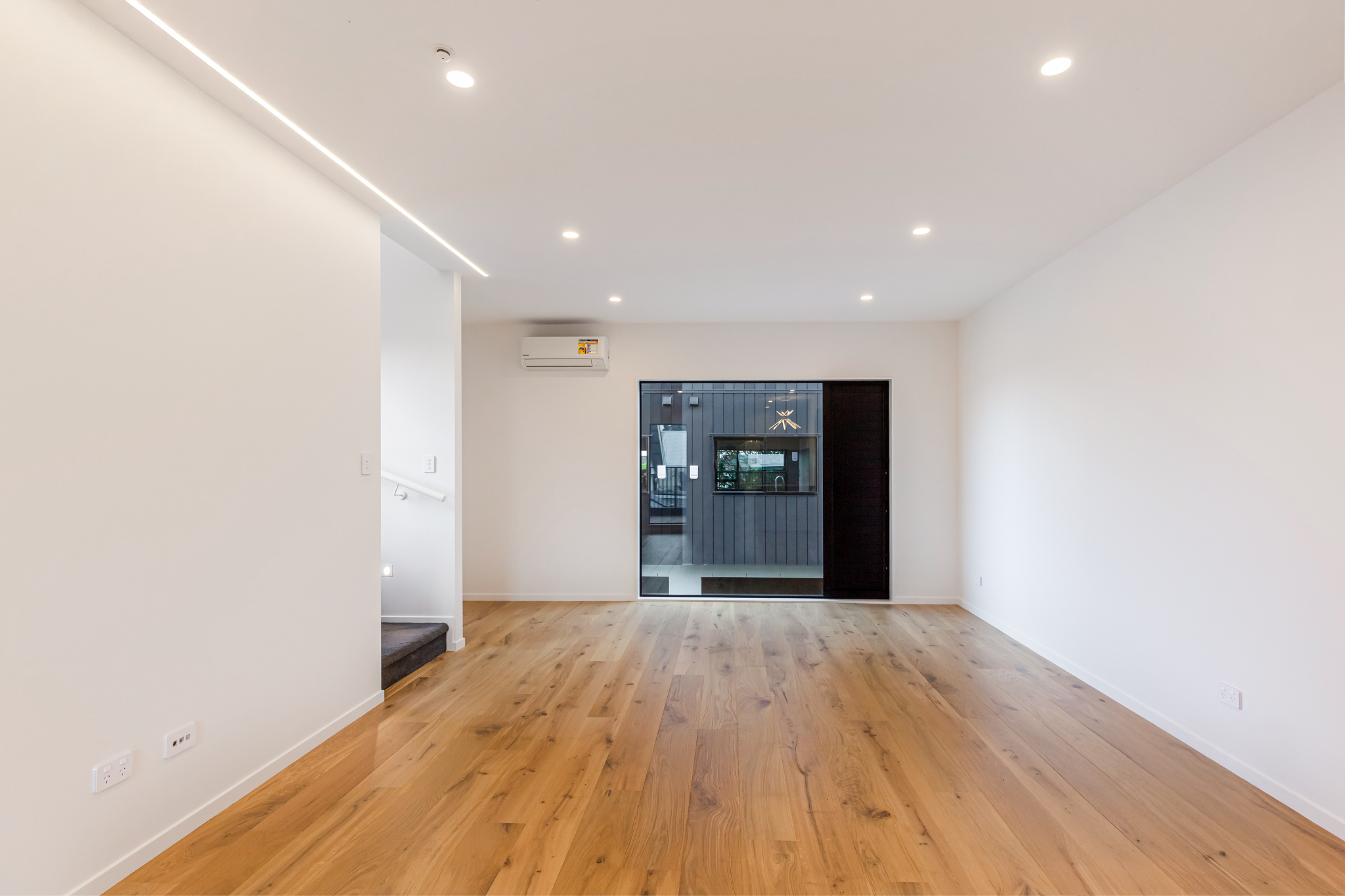 Empty modern living room with white walls, wooden flooring, a black door with glass panel, and an air conditioning unit on the wall.