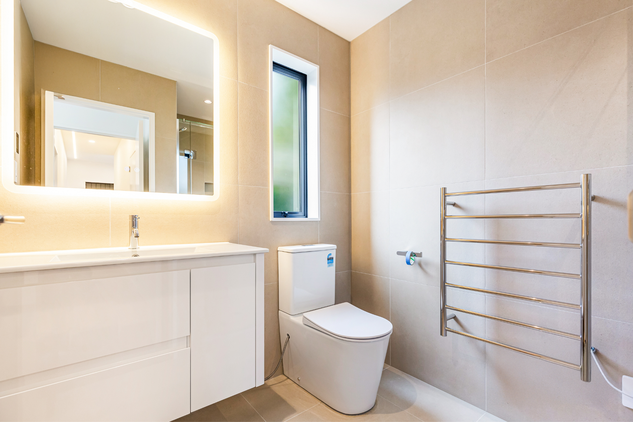 Modern bathroom with beige tiled walls, a white vanity with a sink and a mirror with backlight, a window, a white toilet, and a stainless steel heated towel rack.