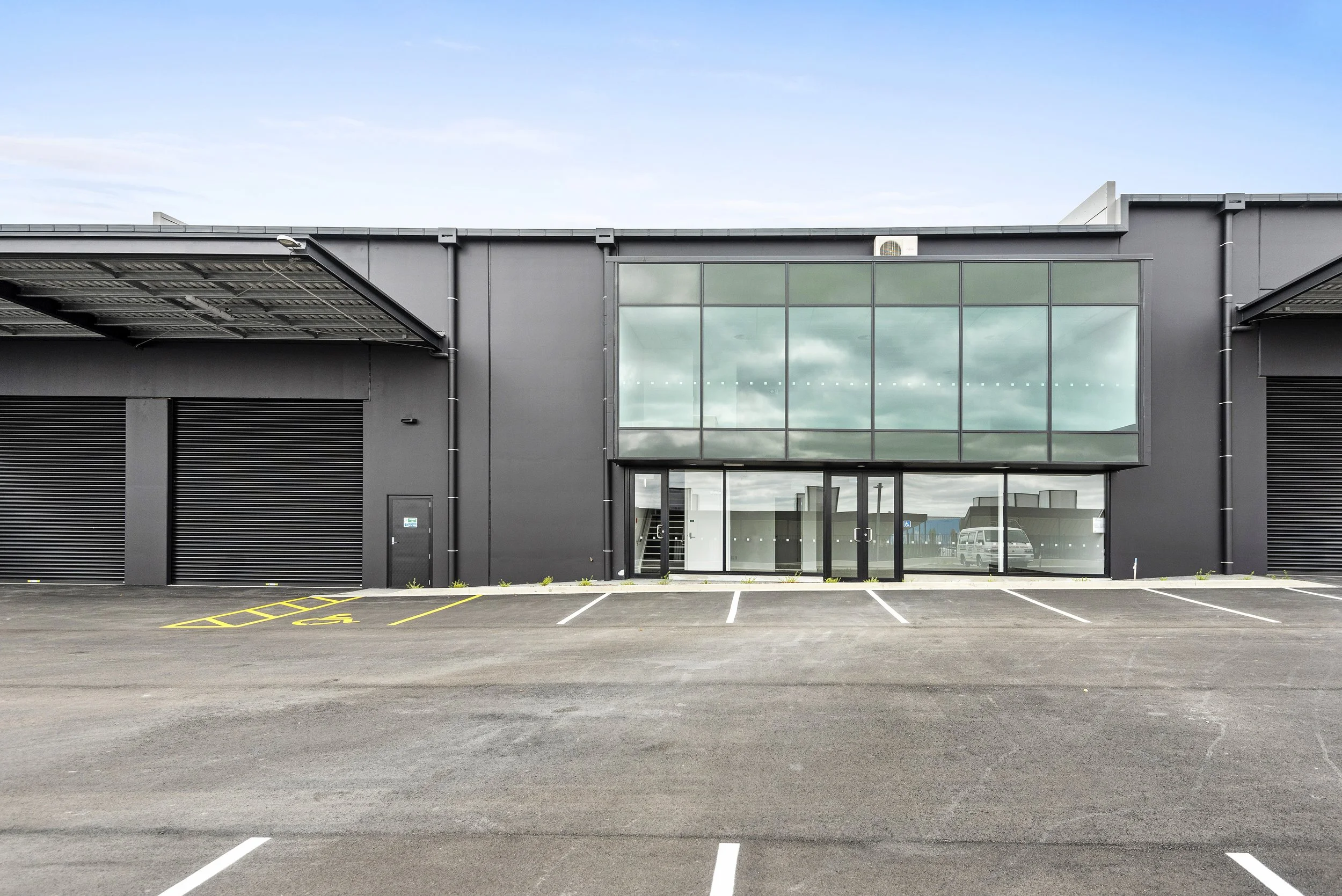 A modern commercial building with large glass windows and black walls, featuring roller shutters on the sides and a parking lot in front with yellow parking space markings.