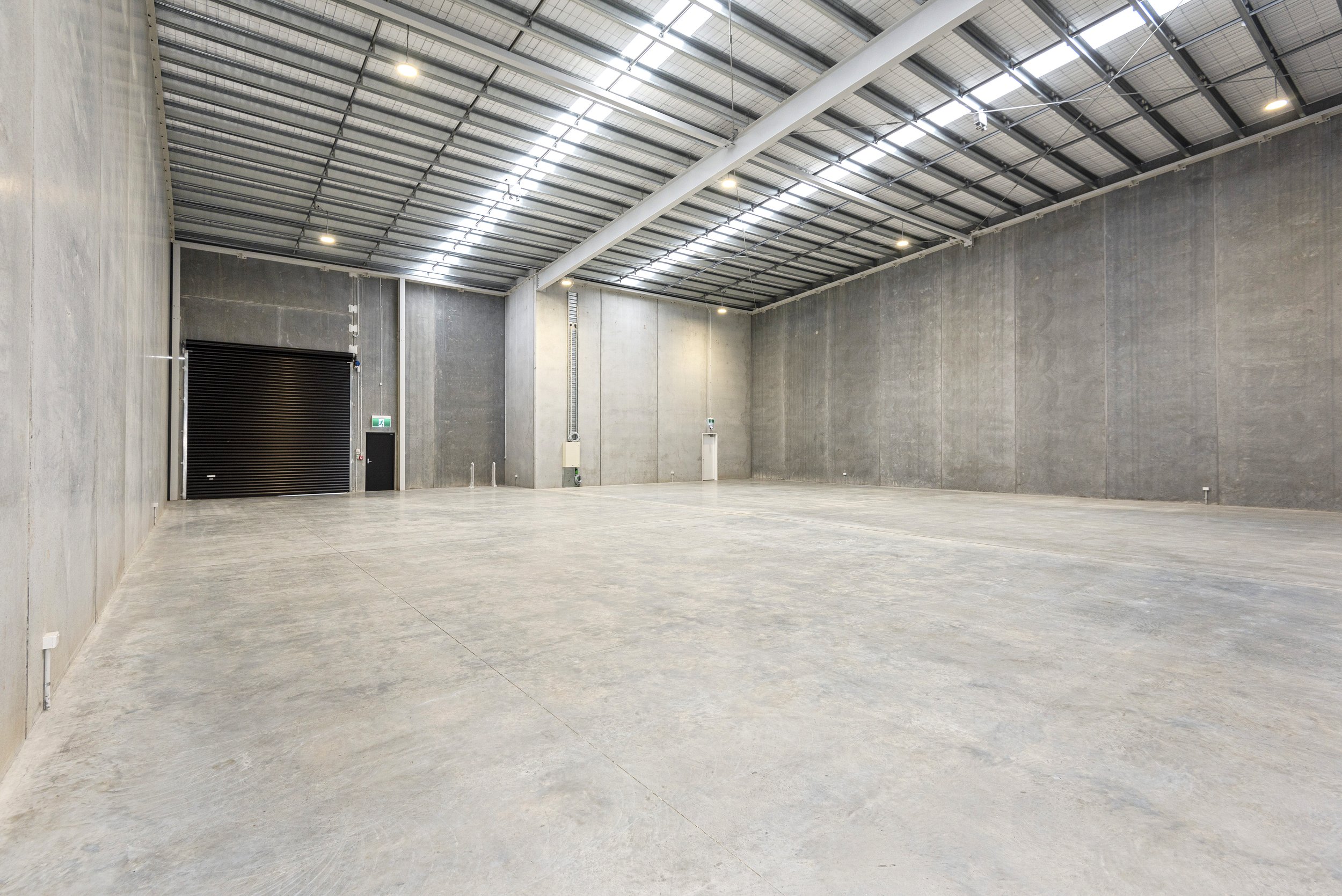 Empty industrial warehouse with concrete walls, polished concrete floor, high ceiling with metal beams, lights, roller door, and utility boxes.