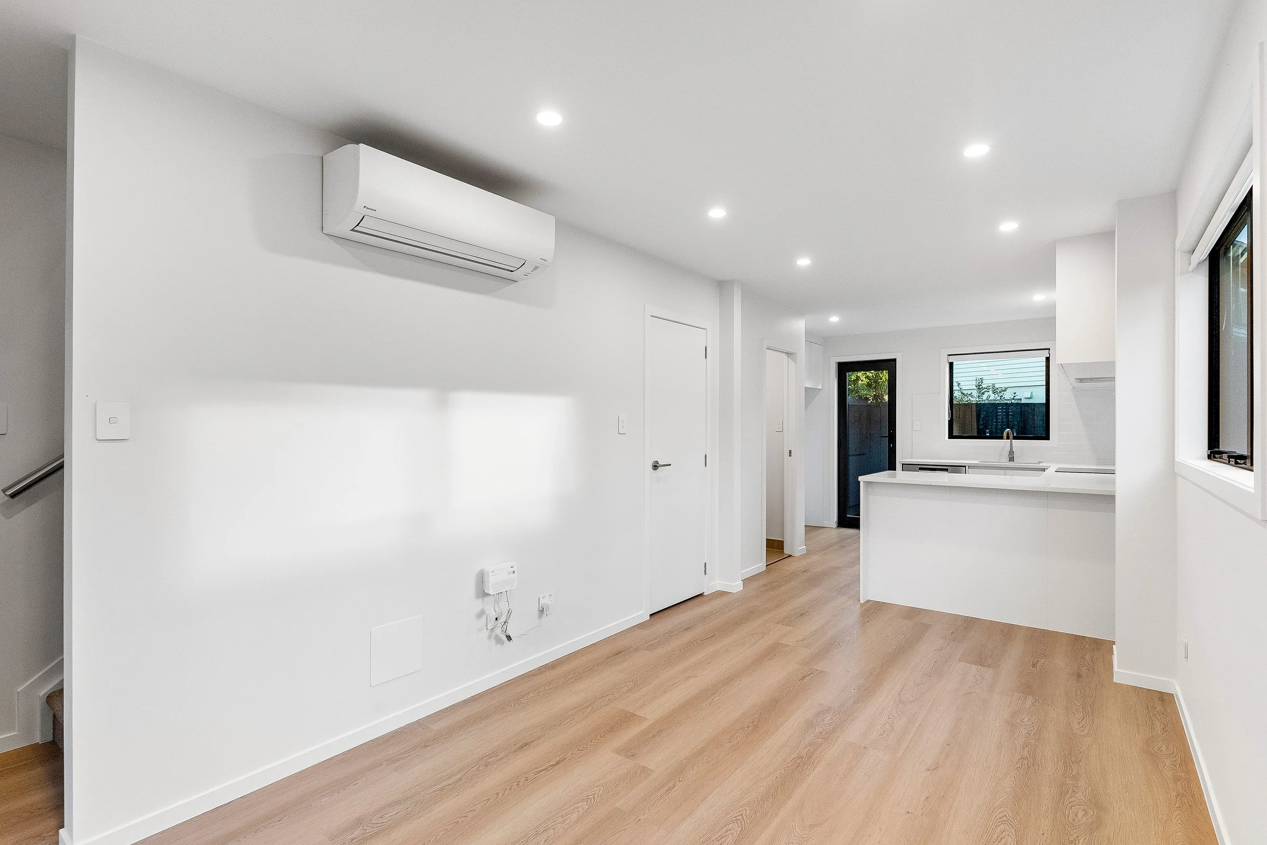 Interior of a modern, minimalistic white kitchen with wood flooring, multiple ceiling lights, large windows, and an air conditioning unit on the wall.