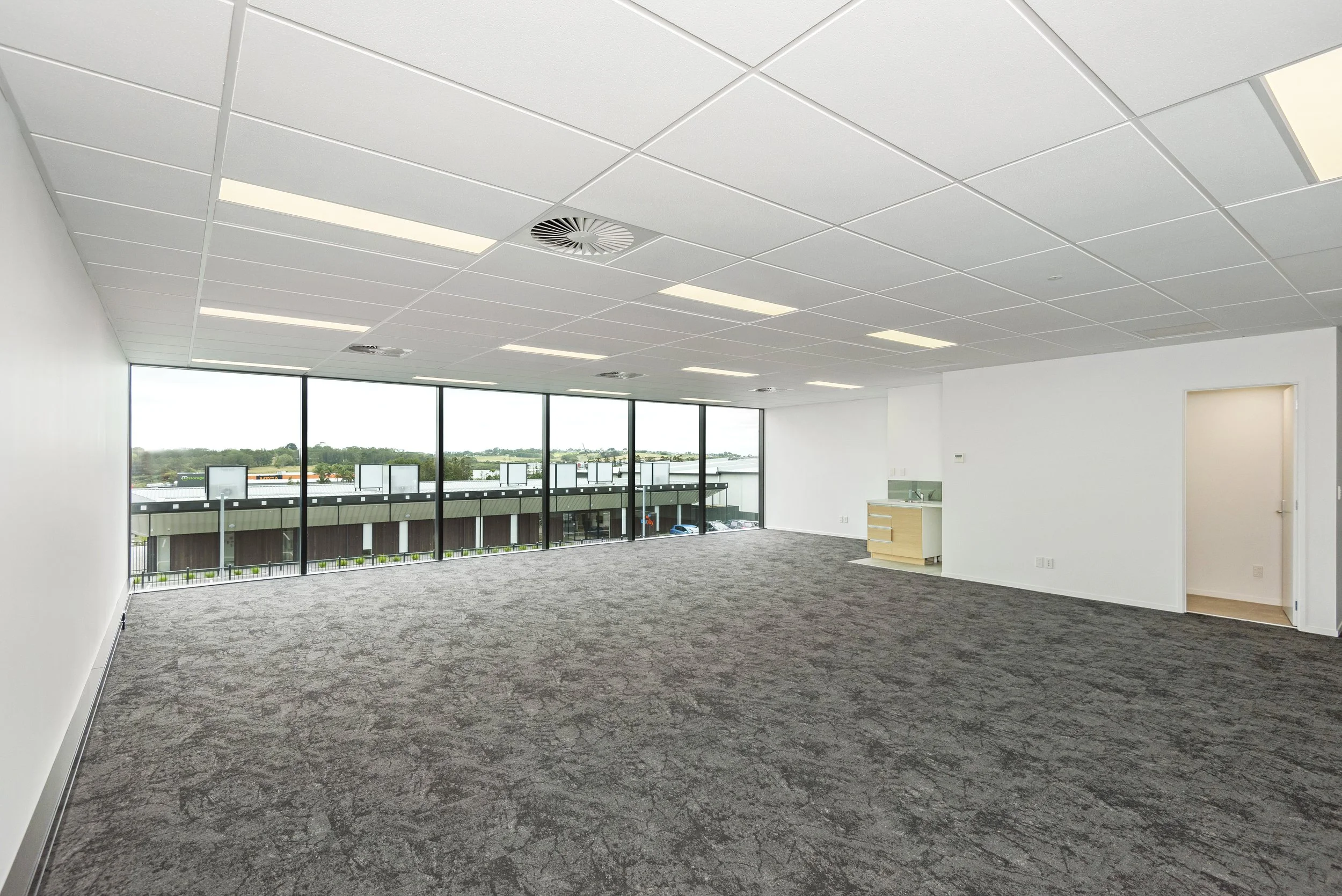 Empty commercial office space with large windows, gray patterned carpet, white walls, and a ceiling with fluorescent lights and air vents.