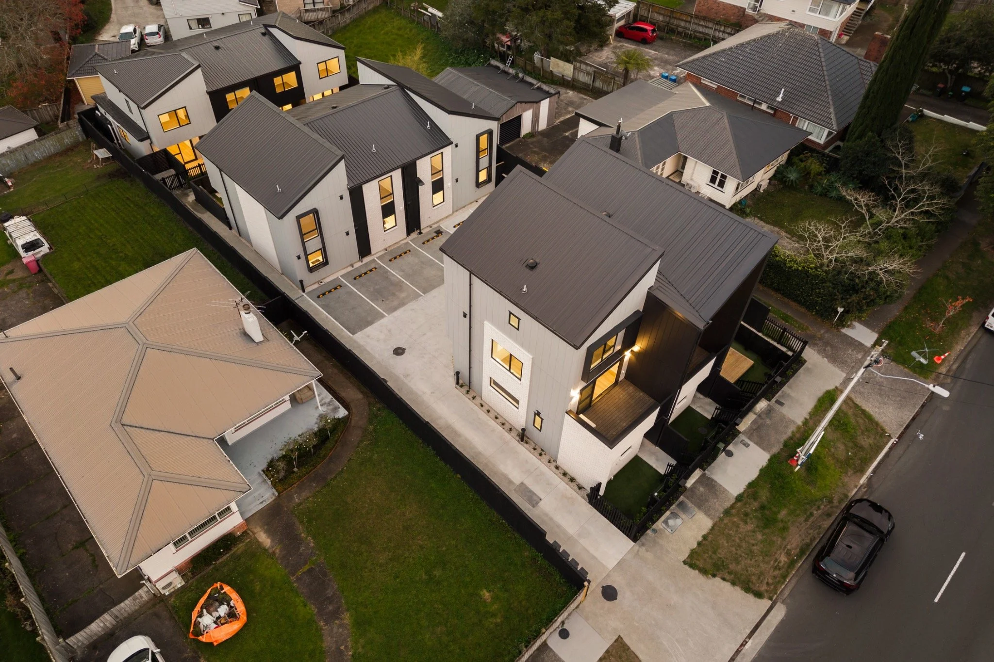 Aerial view of a modern residential building complex with multiple units, black metal rooftops, a parking lot, and surrounding green lawns and neighboring houses.