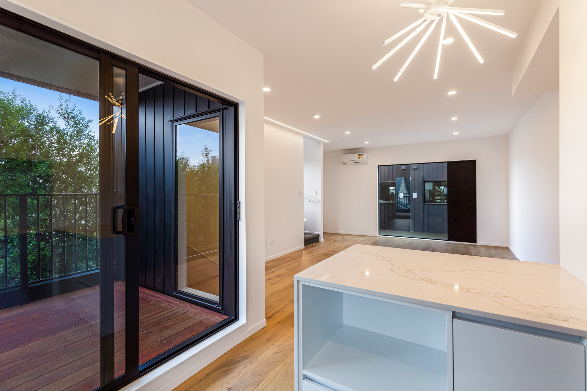 Interior view of a modern, minimalistic room with white walls, wood flooring, a marble countertop island, and a large glass sliding door leading to a balcony with trees outside. Modern ceiling lighting. A black door and window are seen in the background.