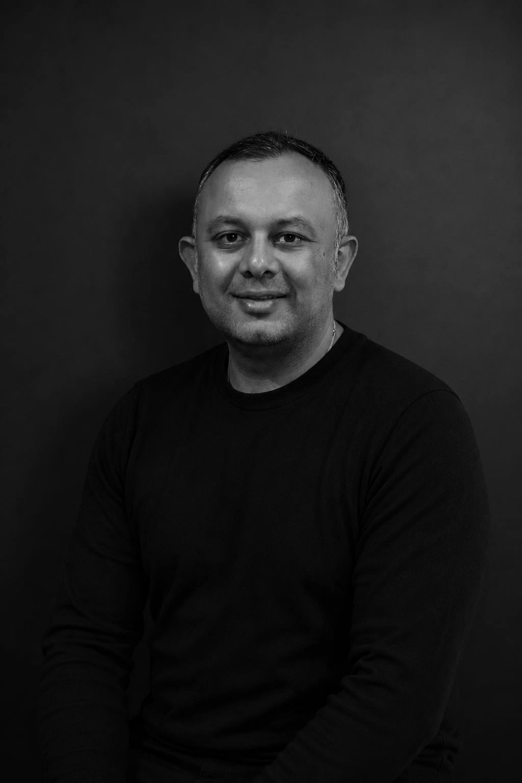 Black and white portrait of a man with short hair, wearing a black shirt, smiling slightly, against a dark background.