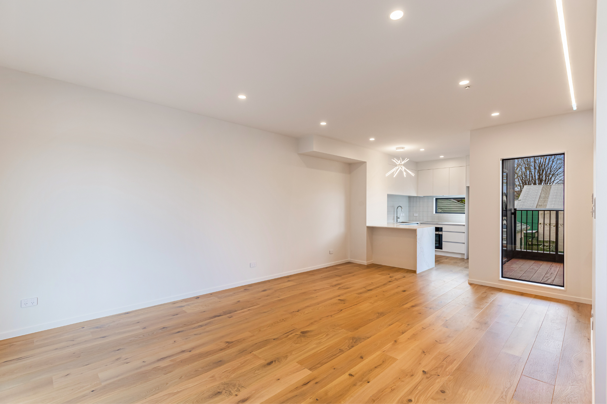 Empty modern living space with white walls, wooden floors, and a kitchen in the background. There is a large window and a sliding door leading to a balcony.