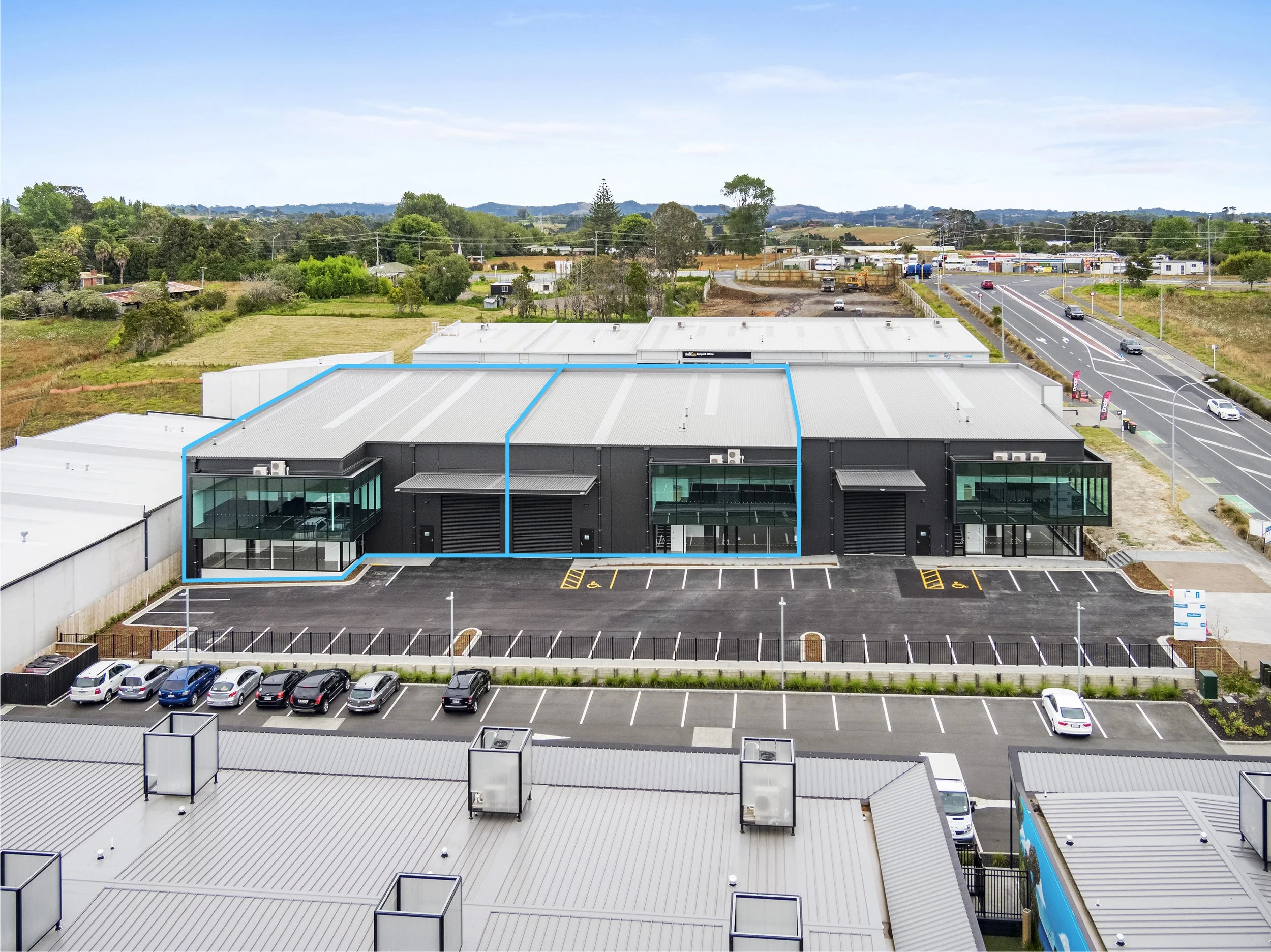 An aerial view of a commercial building with a parking lot, surrounded by green trees, fields, and roads in the background under a partly cloudy sky.