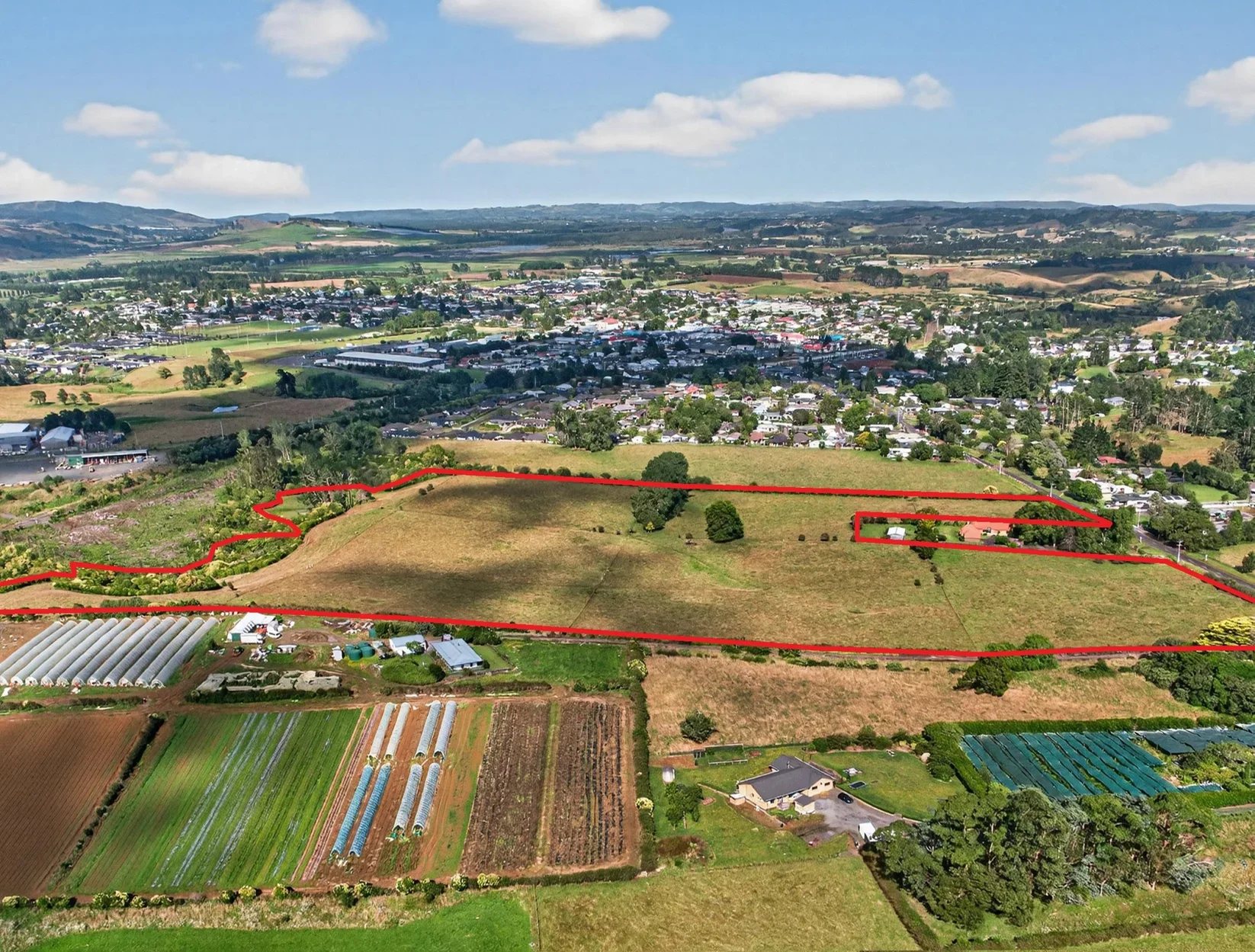 Aerial view of a large plot of land outlined in red, with a small building, trees, and open fields, surrounded by a suburban area and hills in the background.