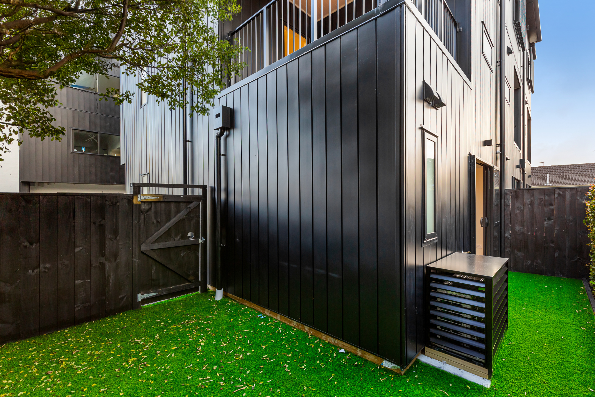 Backyard with black modern building, green artificial grass, and wooden fence. A patio heater is placed on the ground, and a small table is nearby. A tree with green leaves extends into the image from the left side.