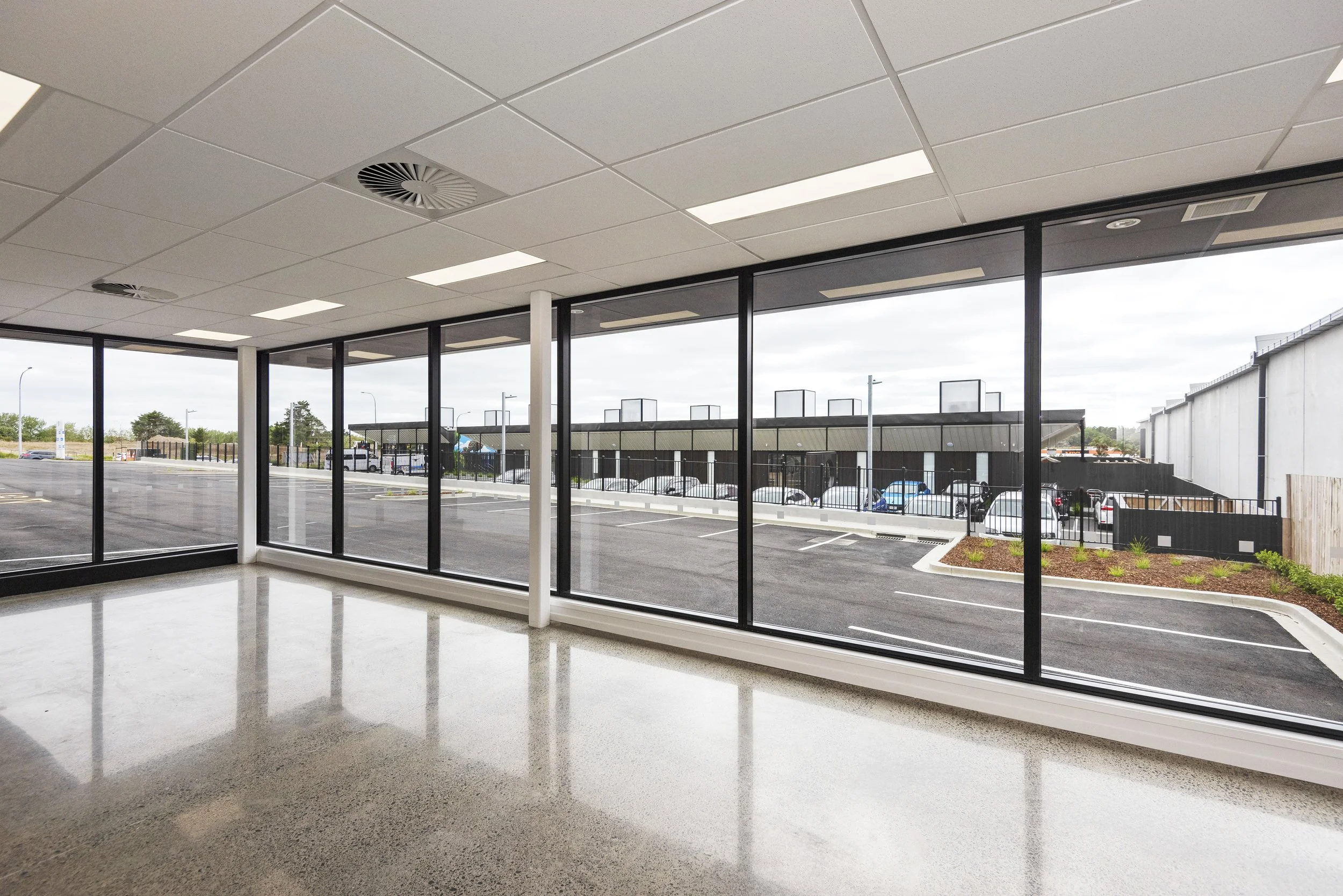 The interior of a commercial building showing large floor-to-ceiling glass windows and doors, with an empty polished concrete floor and a white ceiling with recessed lighting and ventilation vents. Outside, parking lot and surrounding businesses are visible.