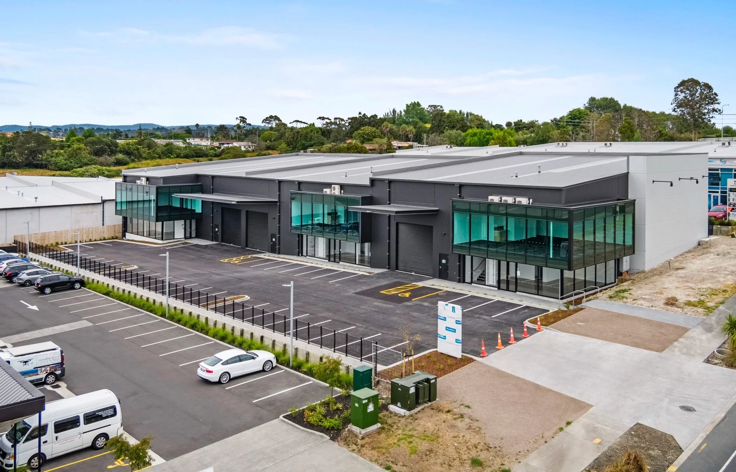 Modern commercial building with glass windows and a parking lot, surrounded by green trees and a blue sky.
