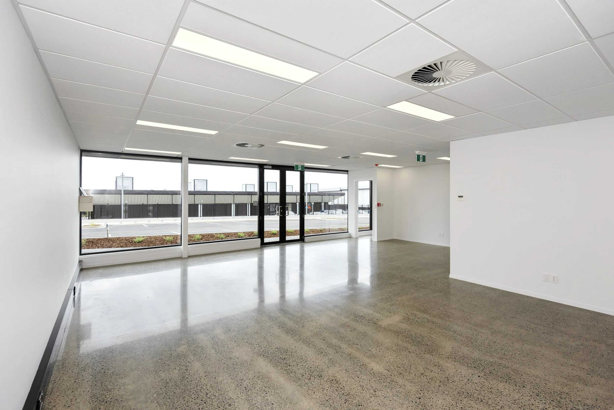 Empty room with white walls, a polished concrete floor, large windows, and a glass door leading outside. The ceiling has rectangular fluorescent lights and vents.
