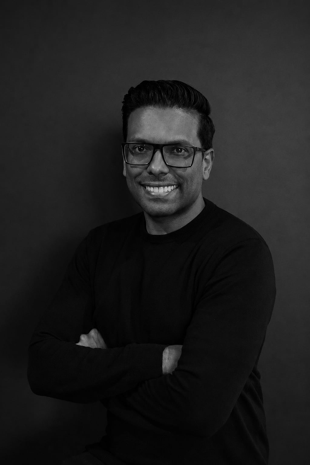 Black and white portrait of a smiling man with glasses, crossed arms, wearing a dark sweater, against a dark background.