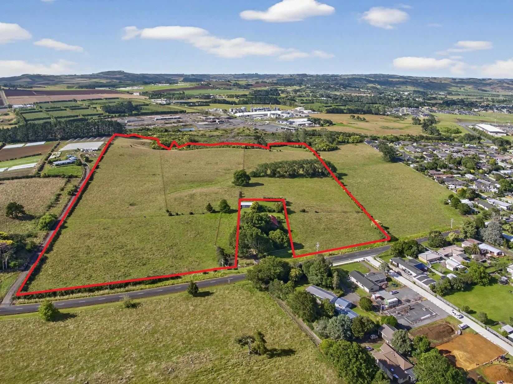 Aerial view of a large green property with a small house and trees, outlined with a red boundary, surrounded by open fields, residential houses, and a distant commercial or industrial area under a partly cloudy sky.