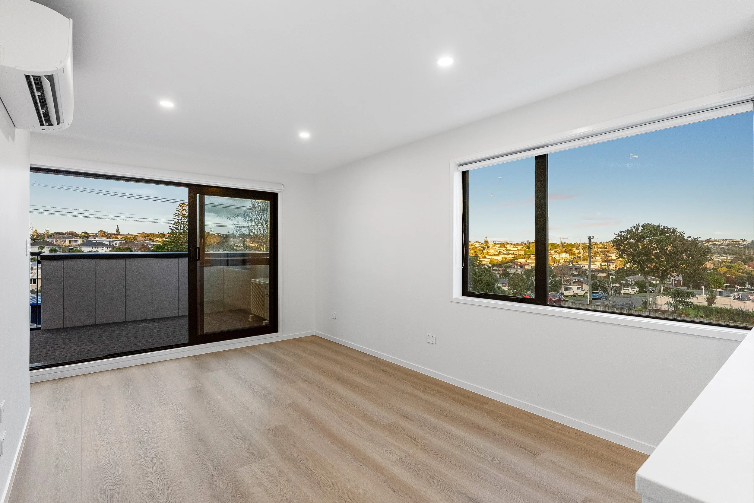 Empty room with wooden flooring, white walls, large windows, and a sliding glass door leading to a balcony with city view.