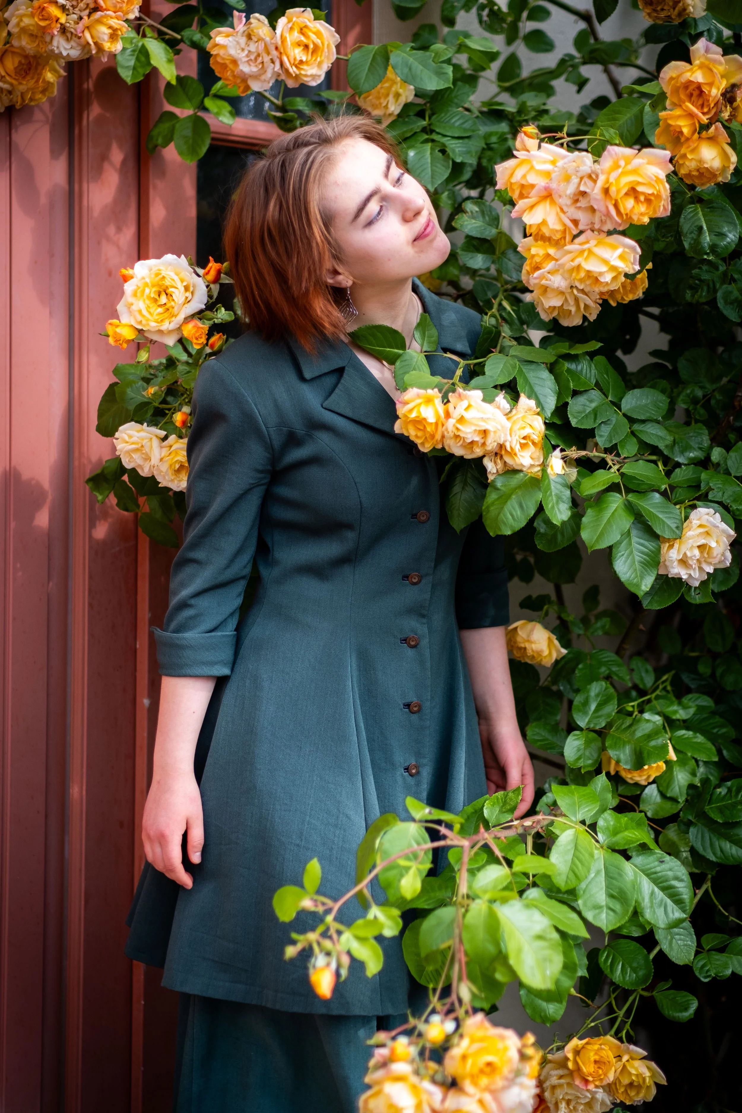 Eine Frau mit roten Haaren steht vor einem Blumenstrauch aus gelben Rosen, umgeben von grünen Blättern.