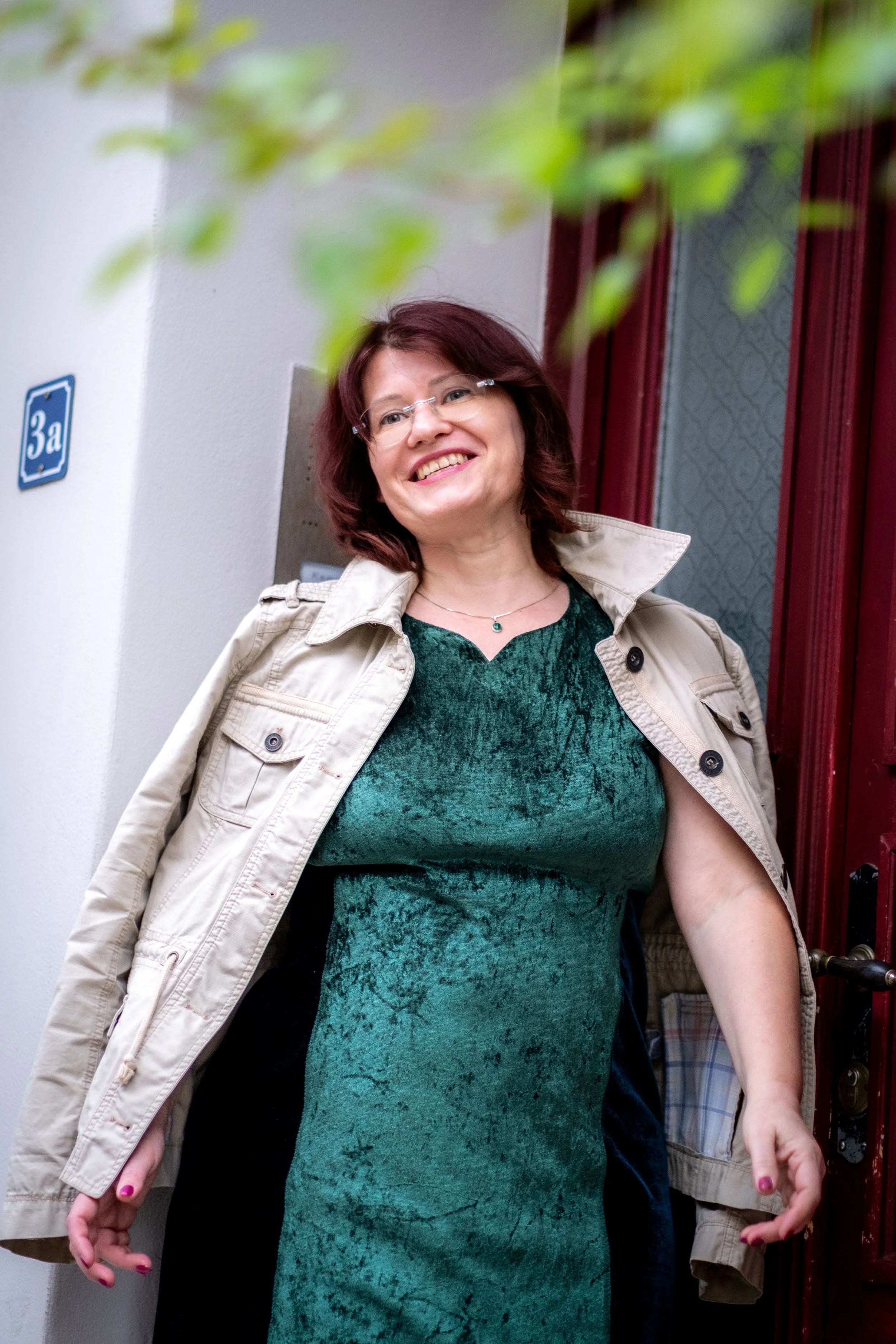 Eine lächelnde Frau mit braunen Haaren, Brille und grüner Kleidung, steht in der Nähe einer Tür. Sie trägt eine beige Jacke und hat auf den Nägeln roten Nagellack.