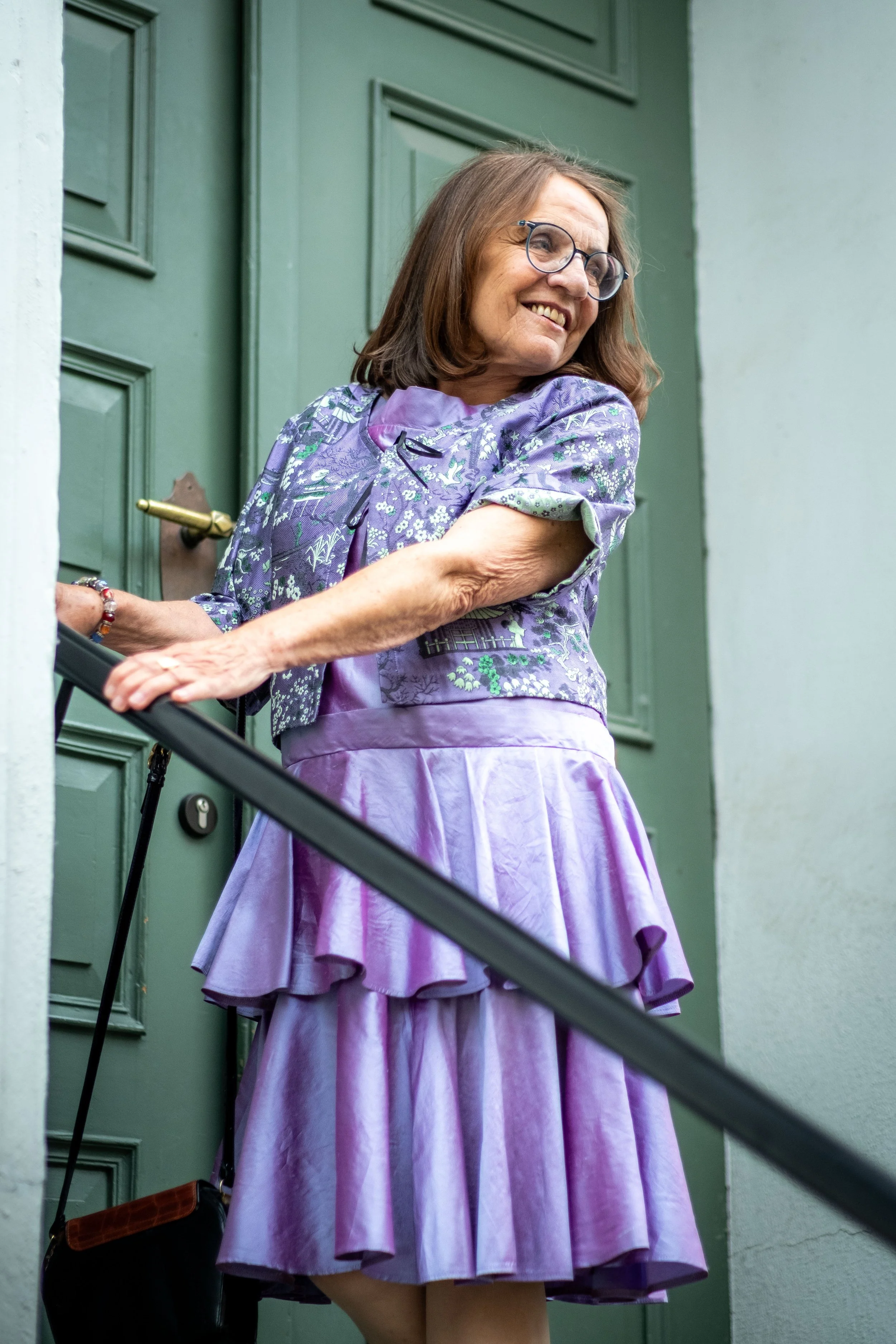 Eine Frau mit Brille in einem lilafarbenen Kleid steht an einem grünen Türrahmen und lächelt.