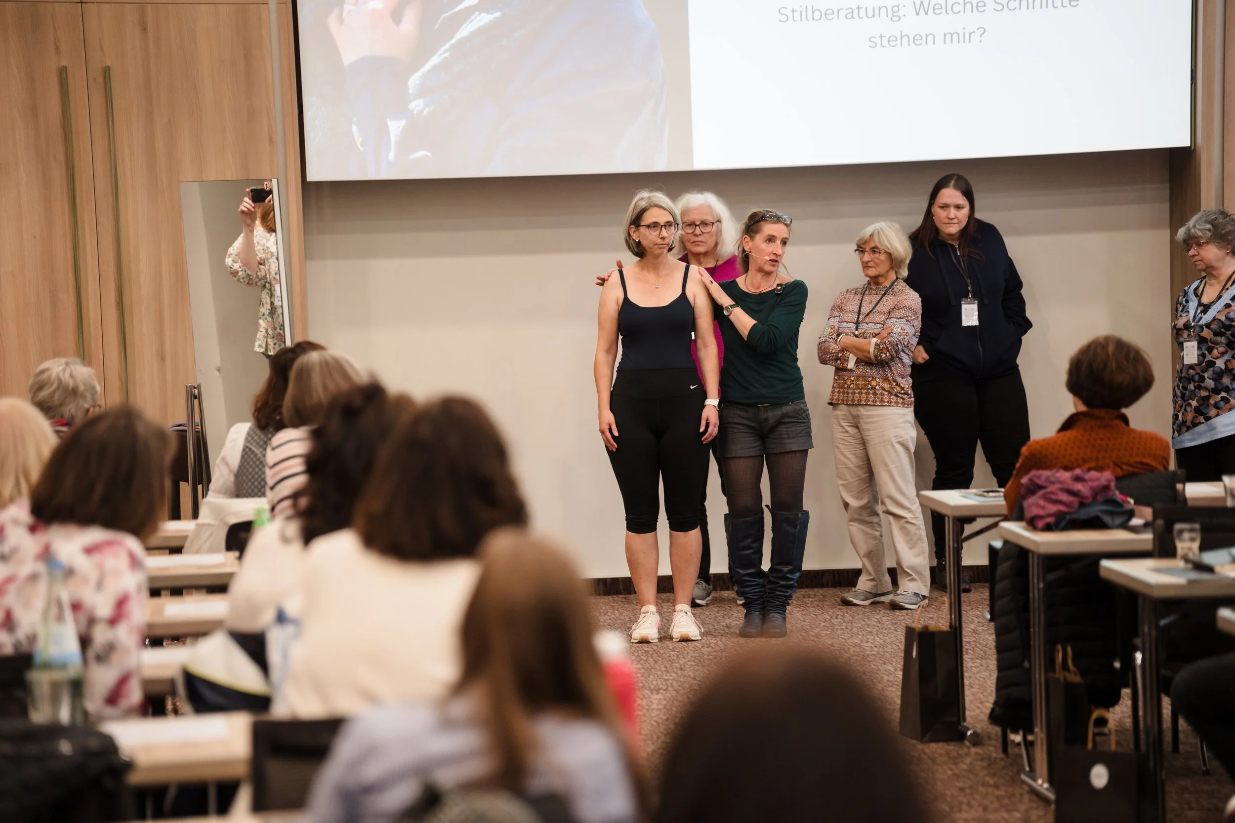 Eine Gruppe von Frauen steht vor einem Publikum in einem Seminarraum. Im Hintergrund ist eine große digitale Tafel mit Text. Die Frauen scheinen an einer Präsentation oder Diskussion teilzunehmen.