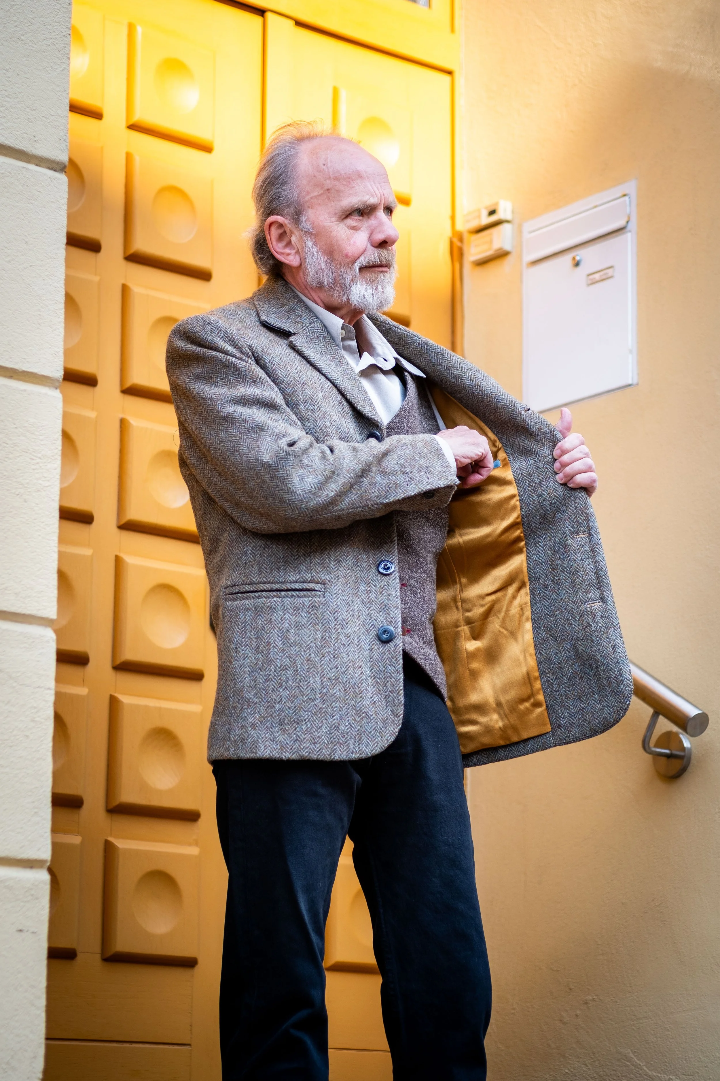 Älterer Mann steht in einem Flur mit gelb-orangefarbenen Wänden und Türverkleidungen, zieht an seinem Jackenärmel und schaut nach rechts.