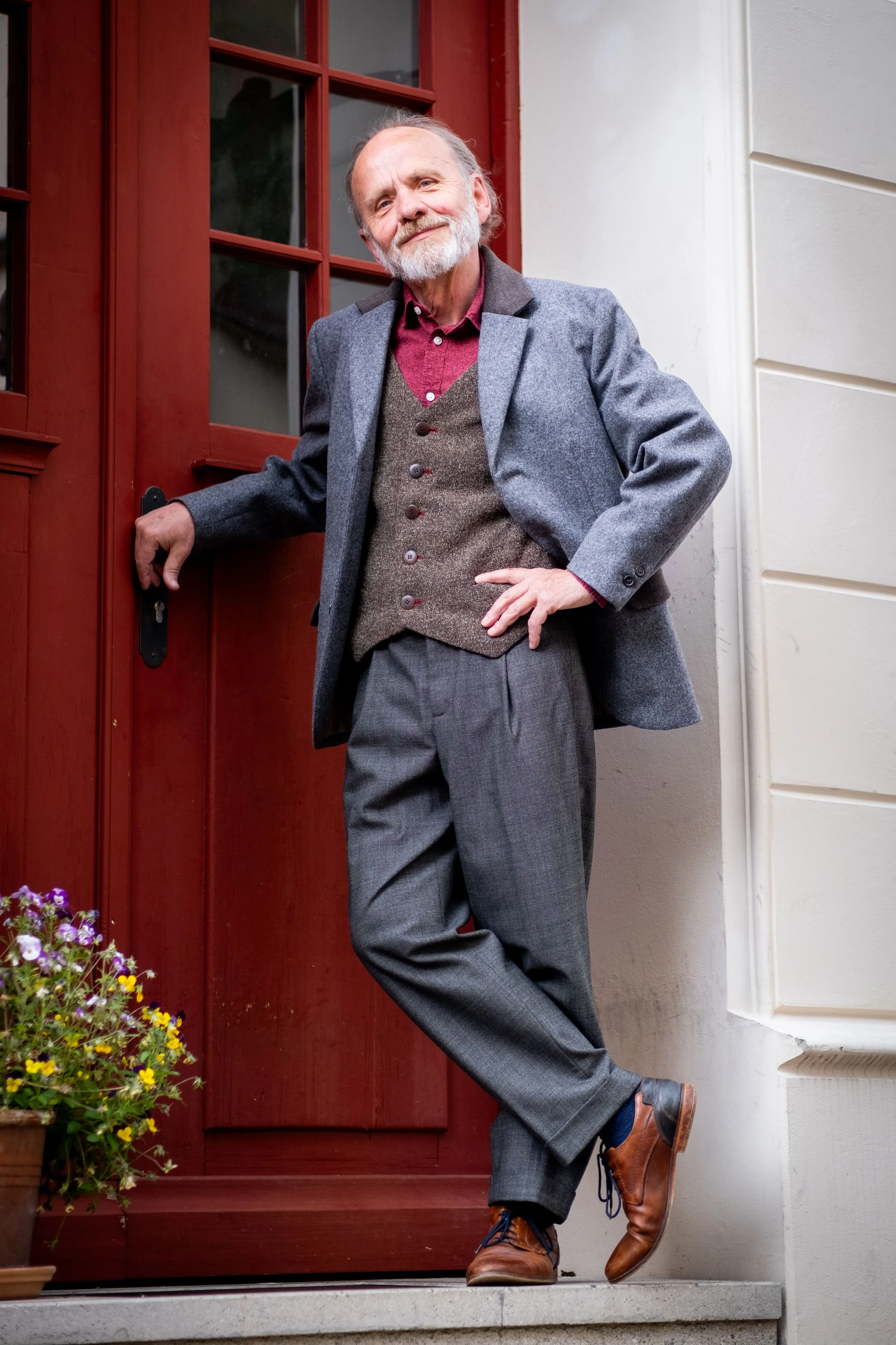 Ein älterer Mann mit grauem Bart und grauen Haaren steht vor einer roten Tür und lächelt. Er trägt einen grauen Mantel, eine braune Weste, eine rot gestreifte Hemd, graue Hosen und braune Schuhe. Er lehnt mit einer Hand an der Tür und hat die andere Hand in der Taille. Im Vordergrund sind Blumen zu sehen.