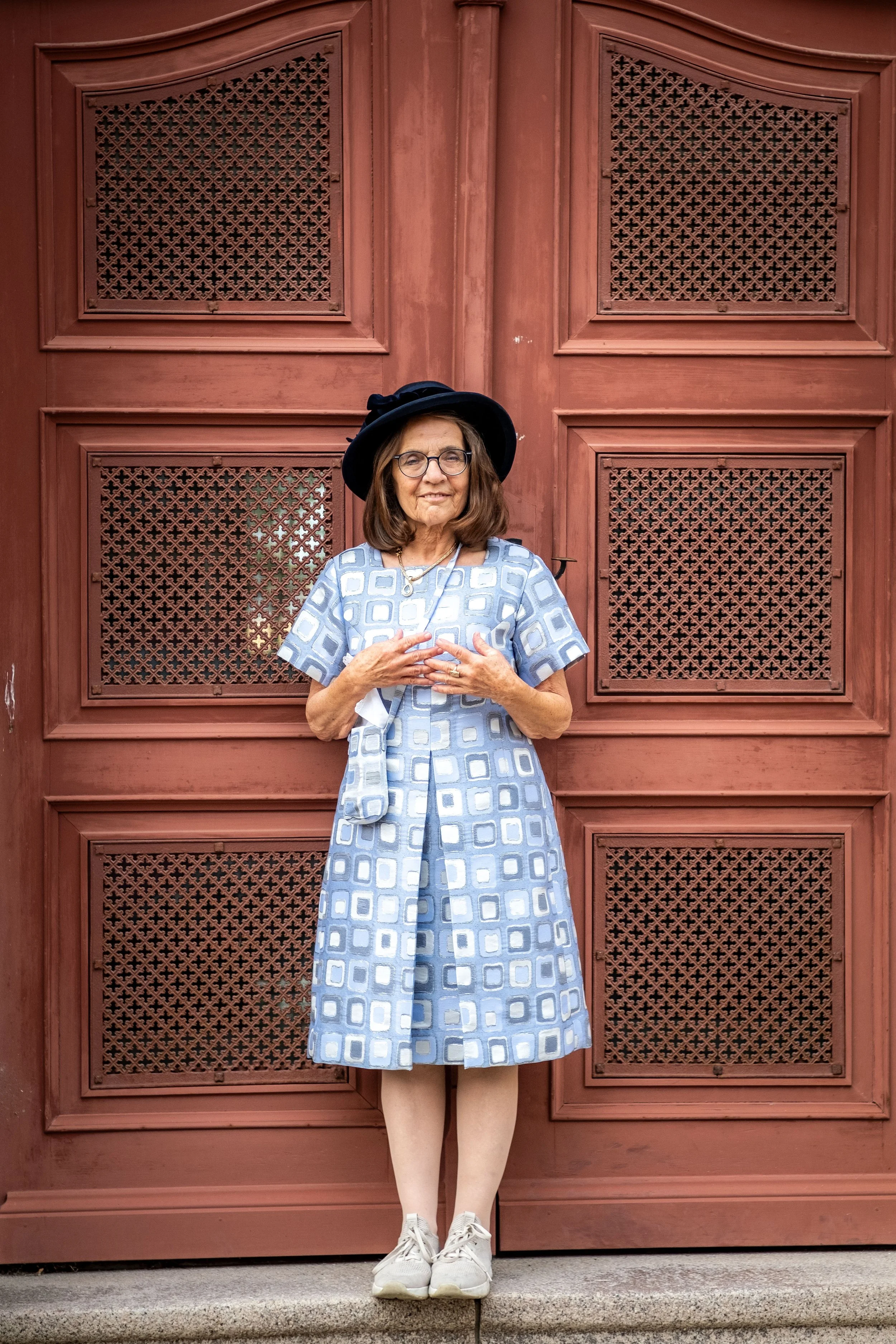 Eine Frau trägt ein blaues, gemustertes Kleid und steht vor einer roten Holztür. Sie trägt einen schwarzen Hut, Brille, eine Halskette und weiße Turnschuhe.