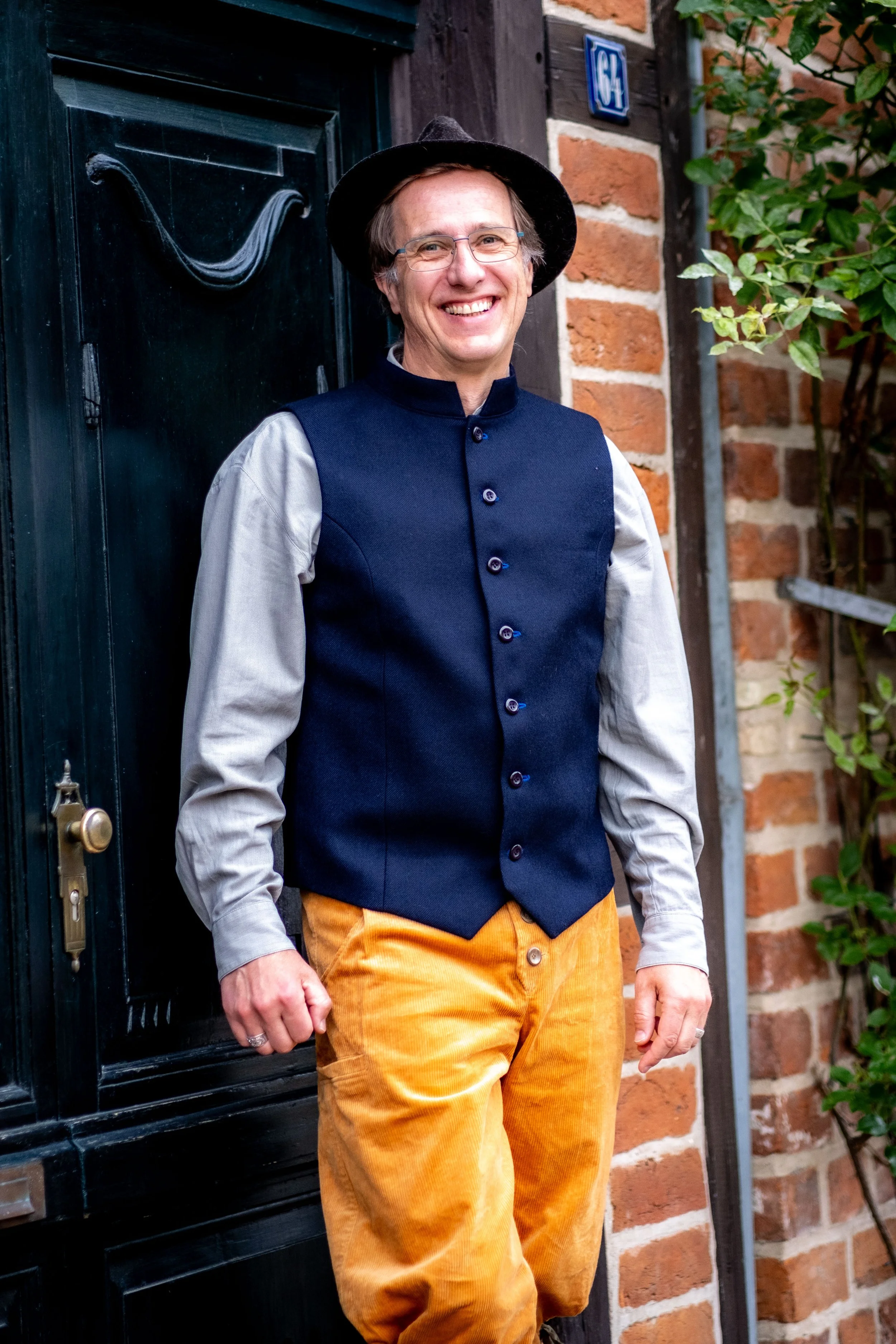 Ein lachender Mann mit Brille in traditioneller Kleidung, bestehend aus einer schwarzen Weste, einem hellgrauen Hemd, orange Latzhose und einem schwarzen Hut, steht vor einer schwarzen Tür und einer Ziegelmauer mit grünen Pflanzen.