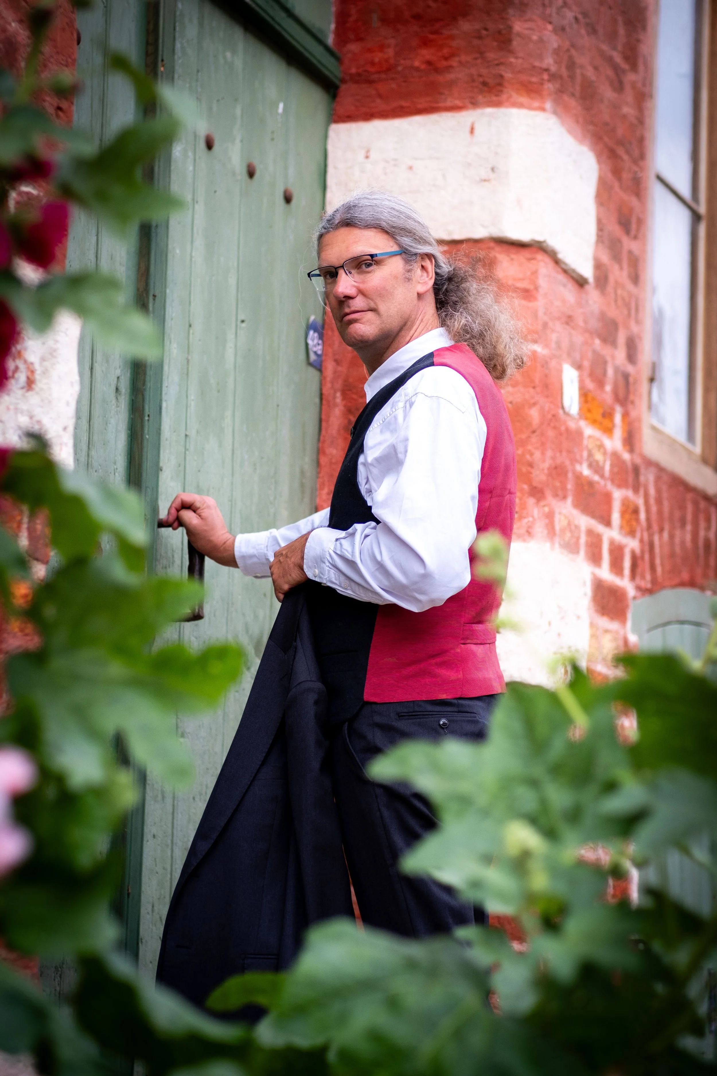 Ein Mann mit grauem, langem Haar, Brille und legerer Kleidung steht vor einer grünen Tür an einem roten Backsteinhaus. Er schaut in die Kamera, während er an der Tür zieht. Vordergrund ist eine grüne Pflanze.