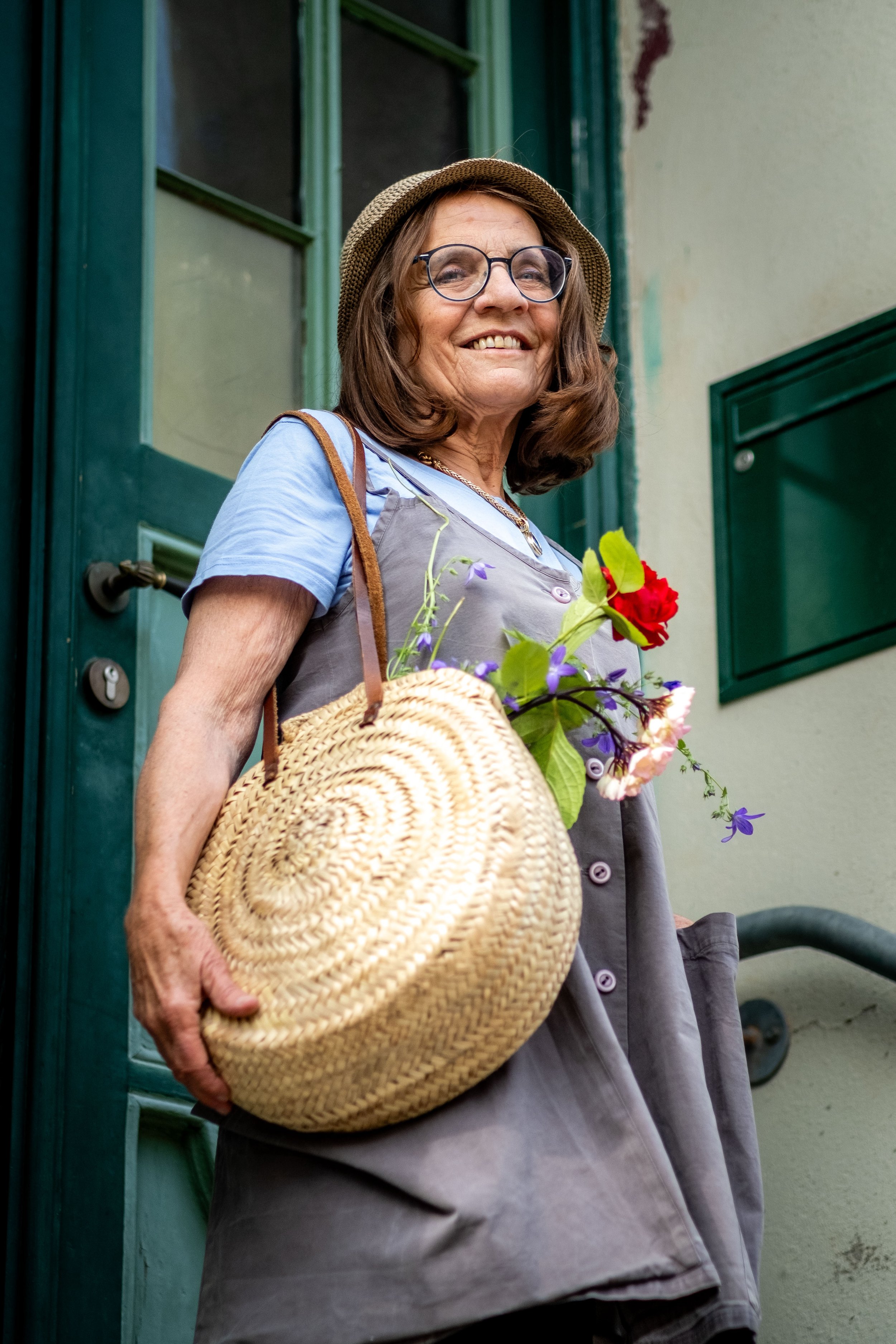 Eine ältere Dame mit Brille, Sonnenhut und einer Tasche mit Blumen steht vor einer grünen Tür und einem Briefkasten und lächelt freundlich in die Kamera.