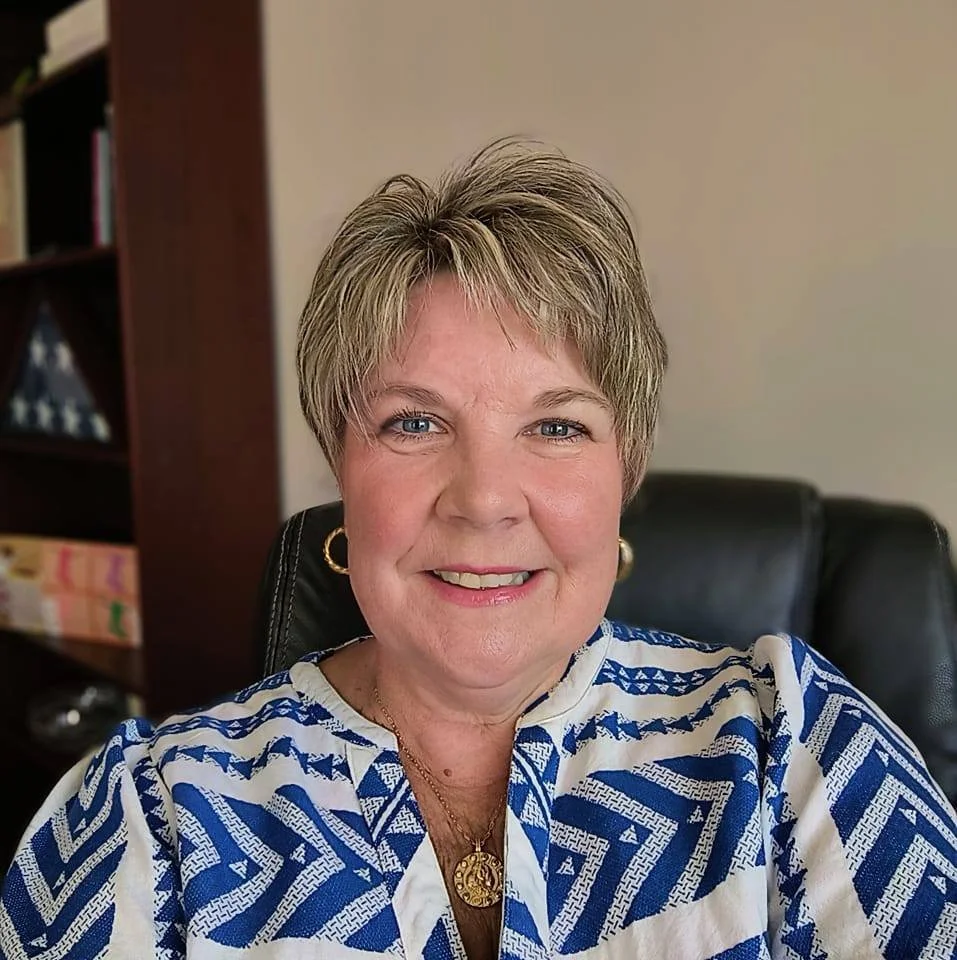 A woman with short blonde hair and blue eyes smiling, wearing a patterned blue and white blouse, sitting on a black office chair in an office with a bookshelf in the background.