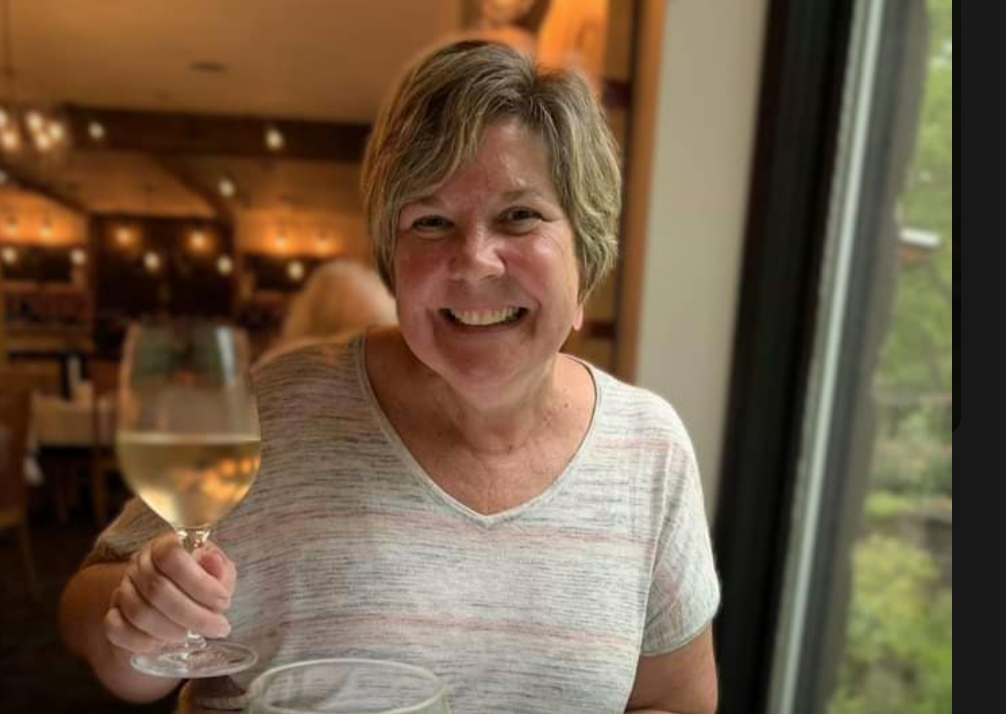 A woman with short hair smiling and holding a glass of white wine in a restaurant with wooden interior and a window showing greenery outside.
