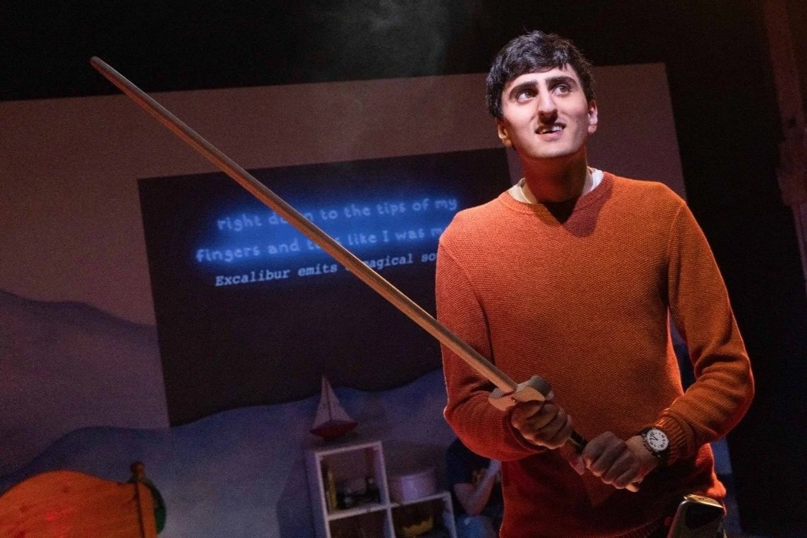 A man with dark hair, wearing an orange sweater and a watch, holding a sword in a room with a screen behind him displaying text, and a bed and shelves visible in the background.