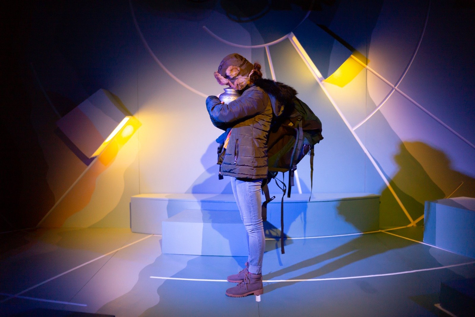 A person with a backpack, dressed in a warm coat, jeans, and boots, stands in a dark room with colorful, abstract light patterns on the walls, seemingly looking at a display or exhibit.