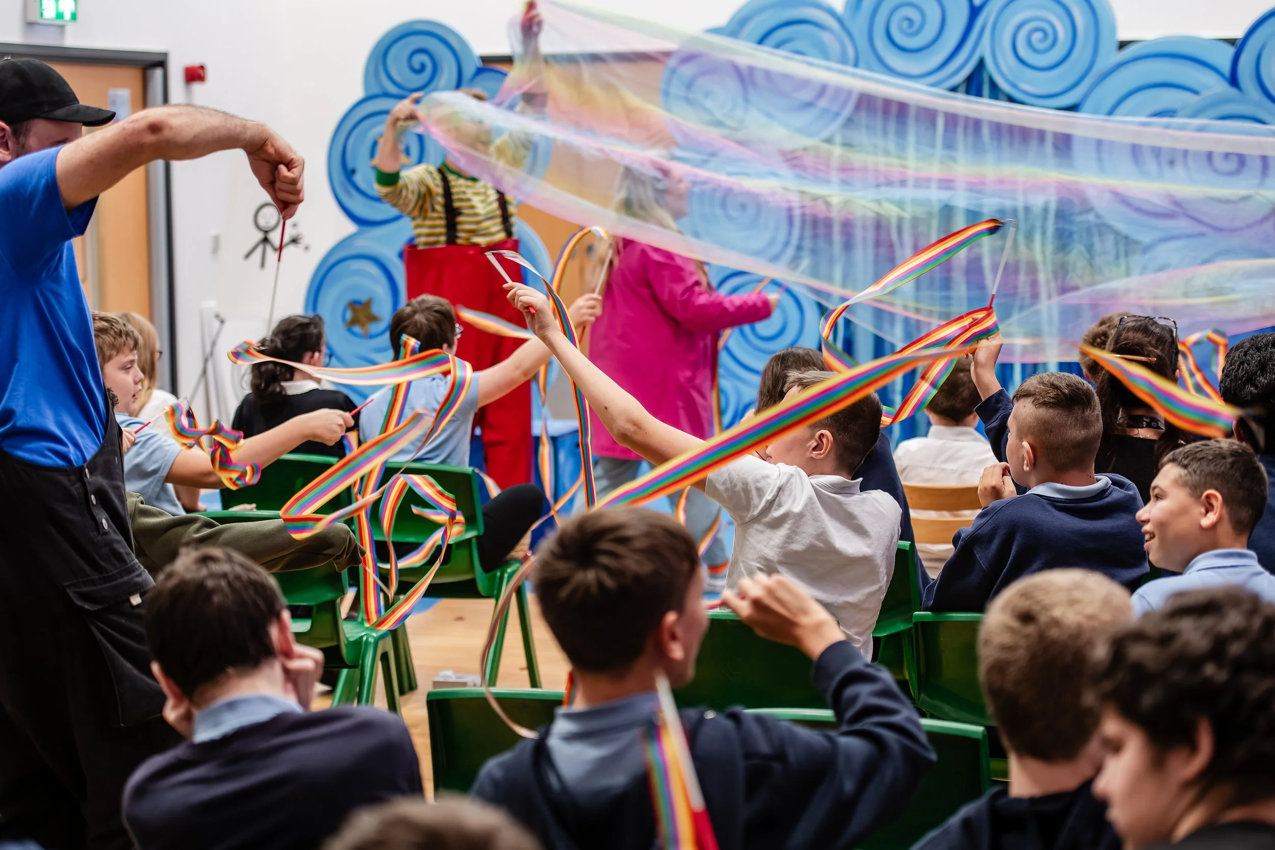 Children and adults participating in a colorful bubble-blowing activity, with rainbow ribbons and a large rainbow soap bubble, against a blue decorative background.